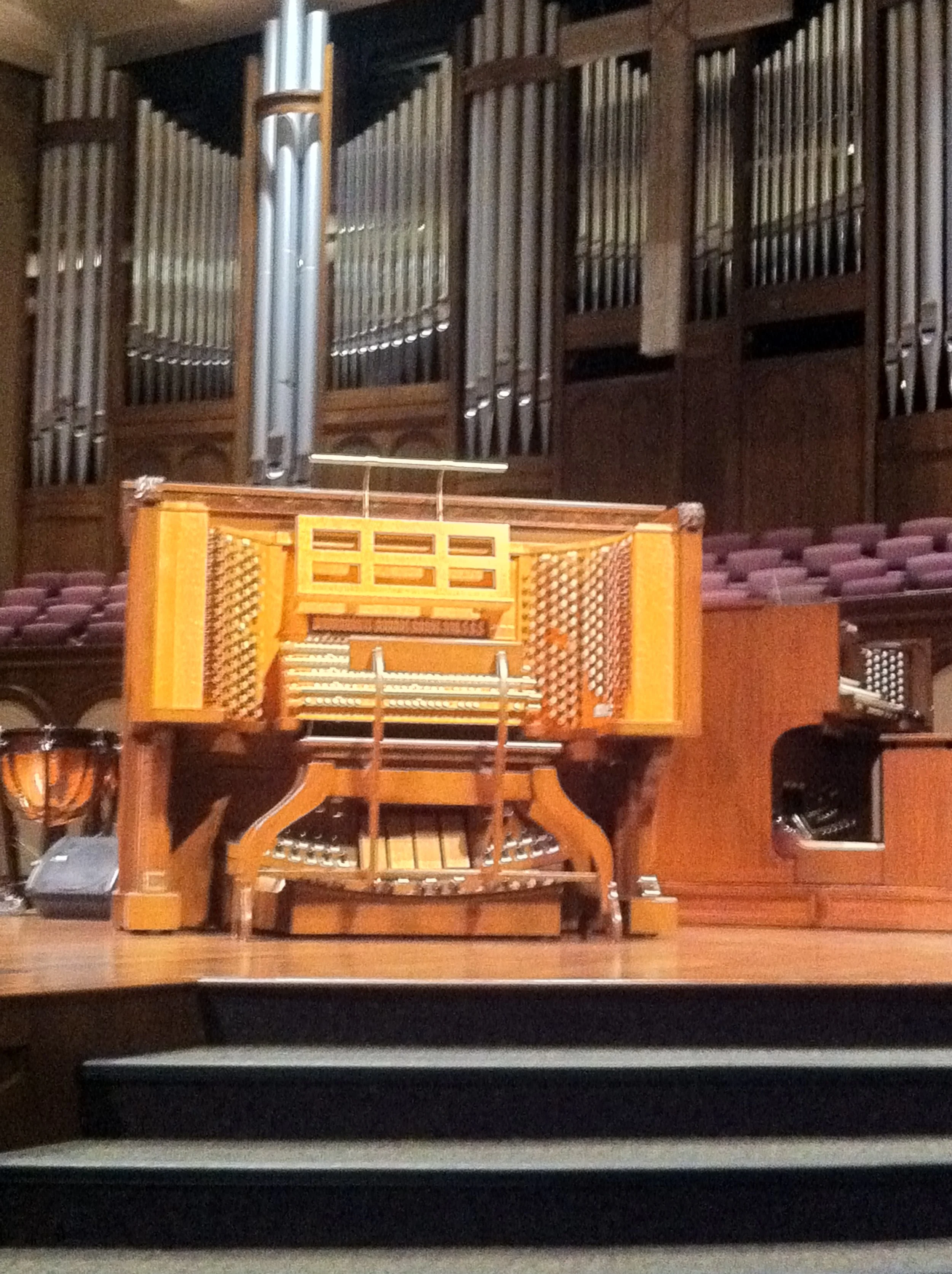 A large organ with pipes, situated in a concert hall or church, with wooden panels and seats visible in the background.