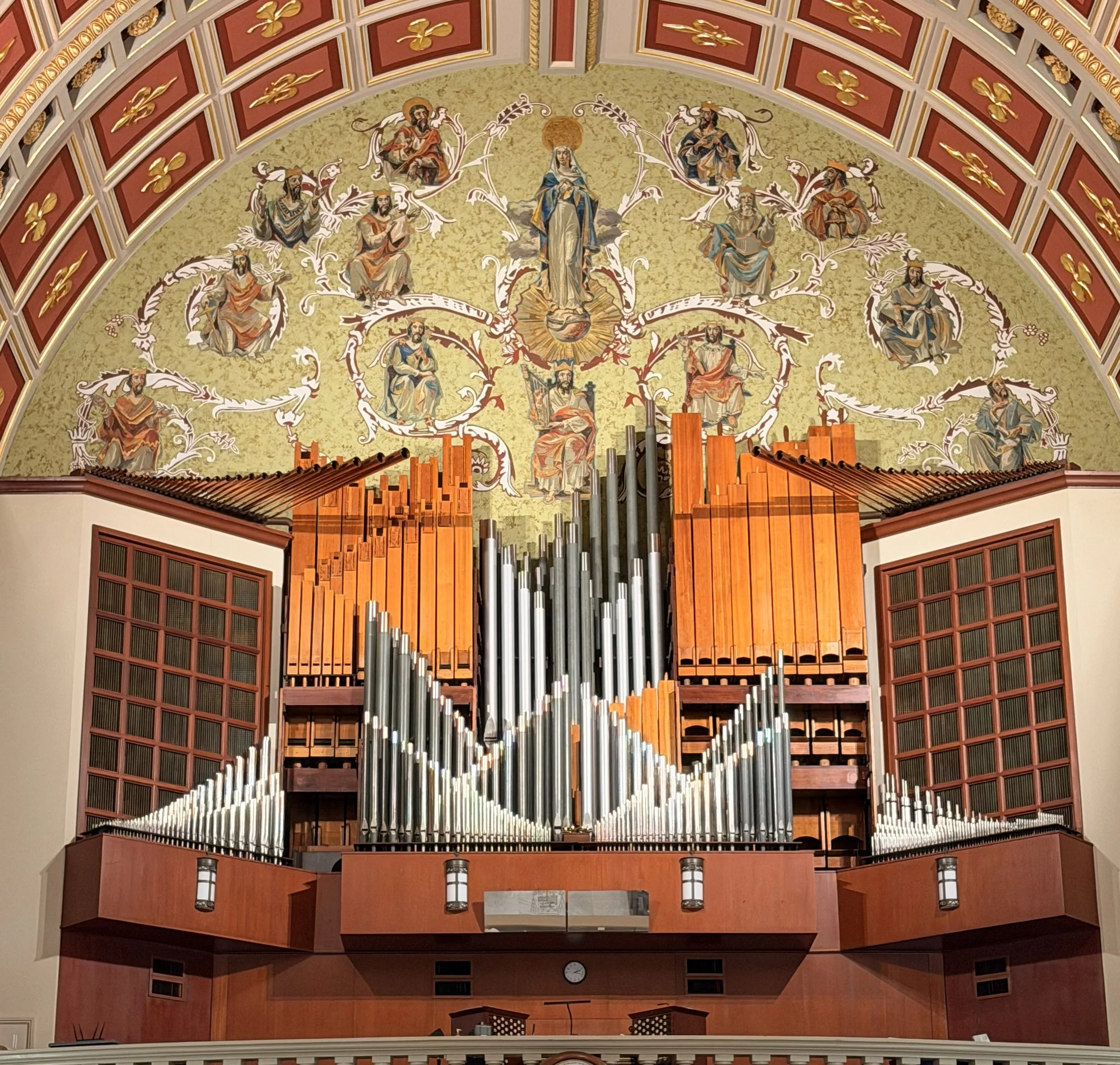 A large pipe organ with orange and silver pipes in a church, with a painted ceiling featuring religious figures and gold detailing.