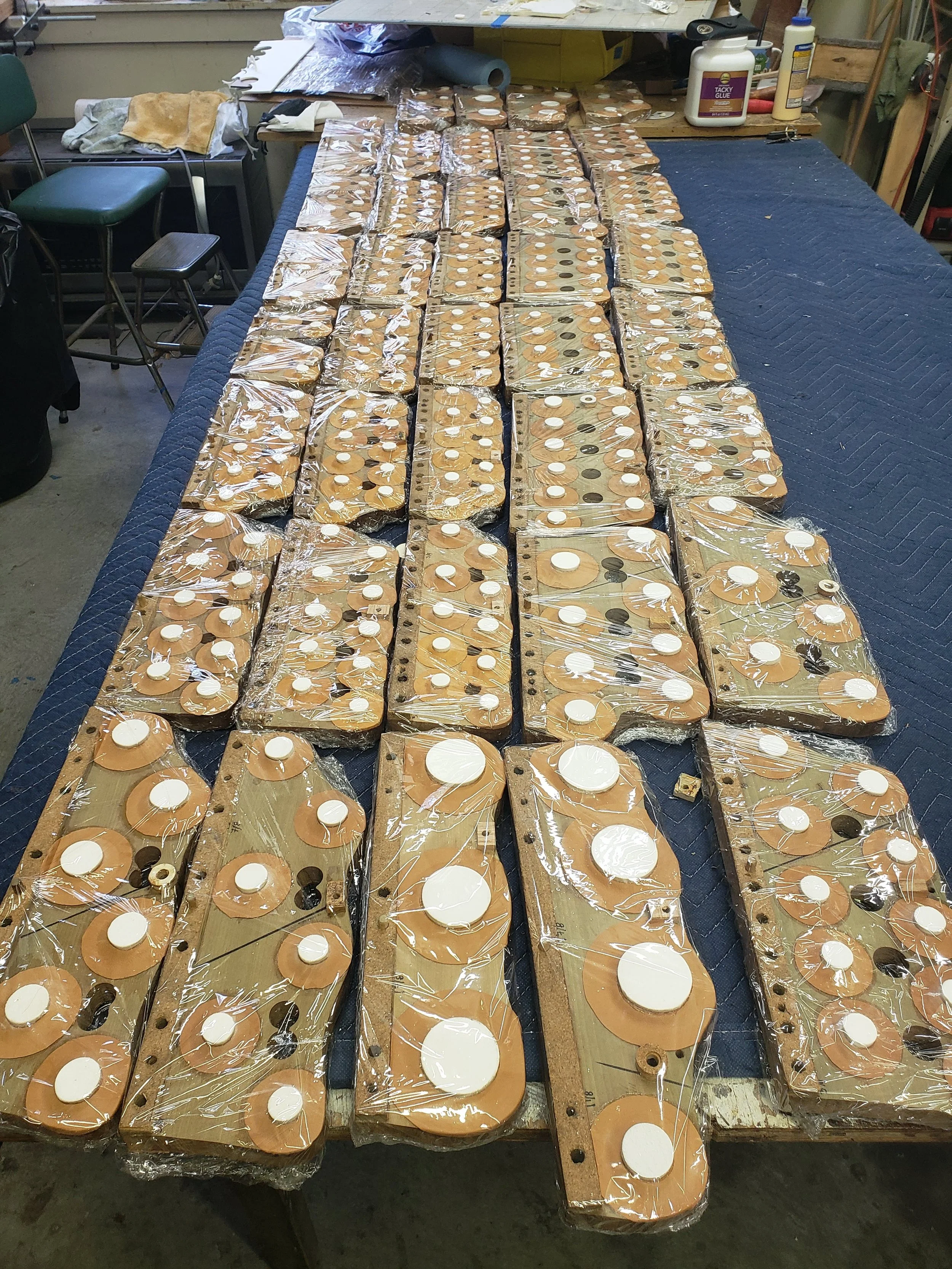 Multiple wooden arcade game controls with circular white, black, and tan buttons, wrapped in clear plastic, laid out on a worktable in a workshop.