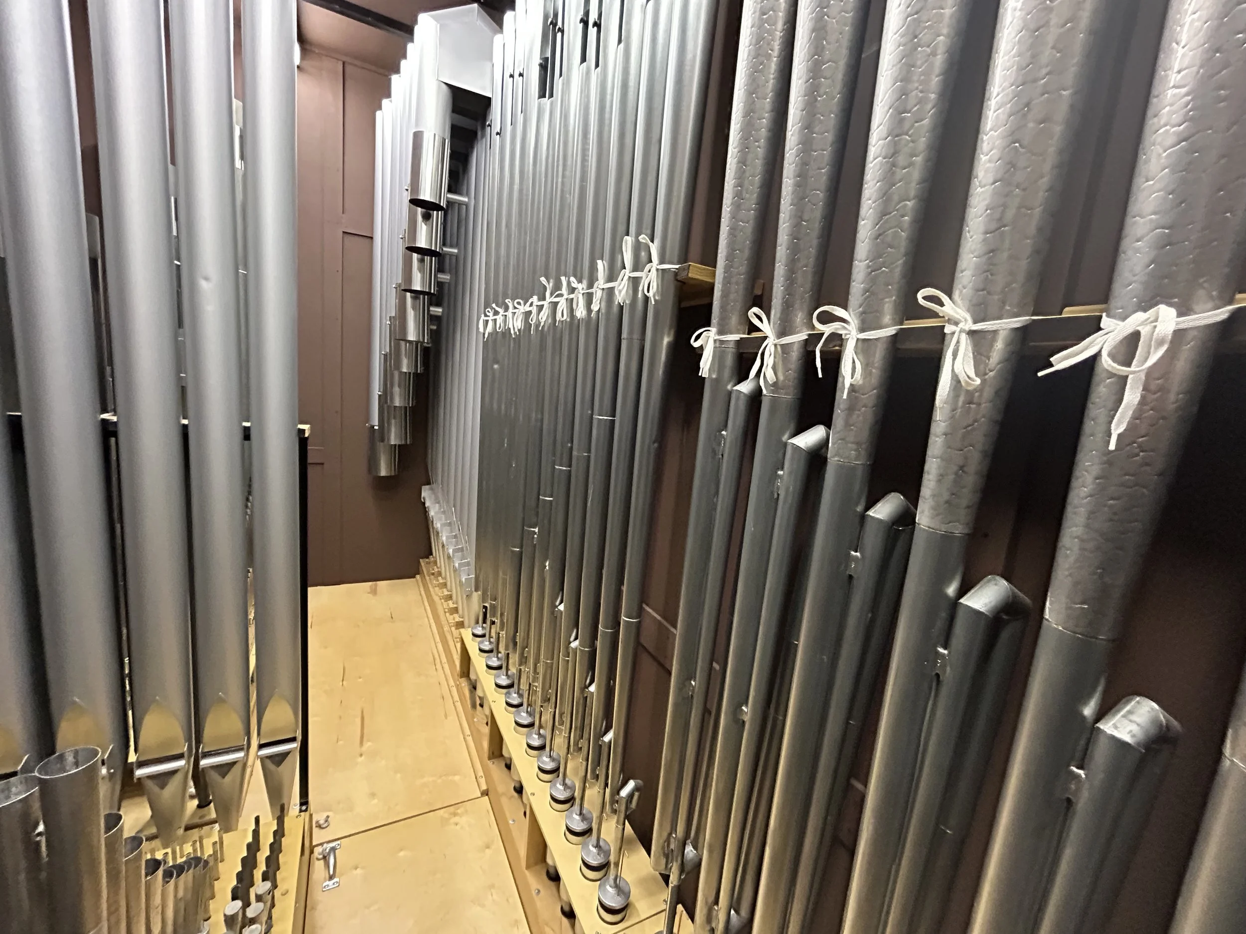 A collection of silver metal pipes stored vertically on a wooden rack, with some tied with white string, inside a storage room.