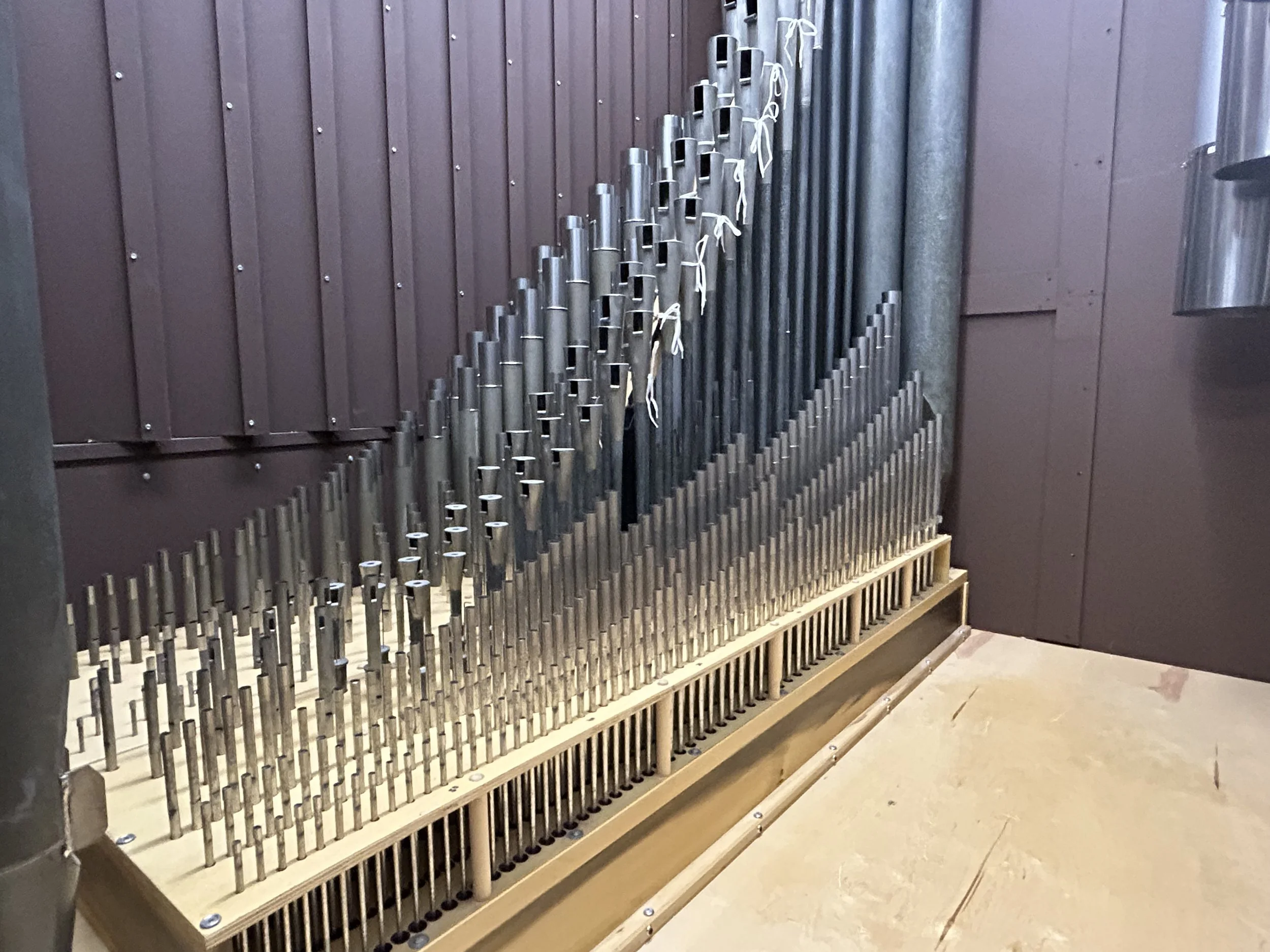 A set of metal pipes arranged in a descending pattern, likely part of a pipe organ, against a brown wall with vertical paneling.