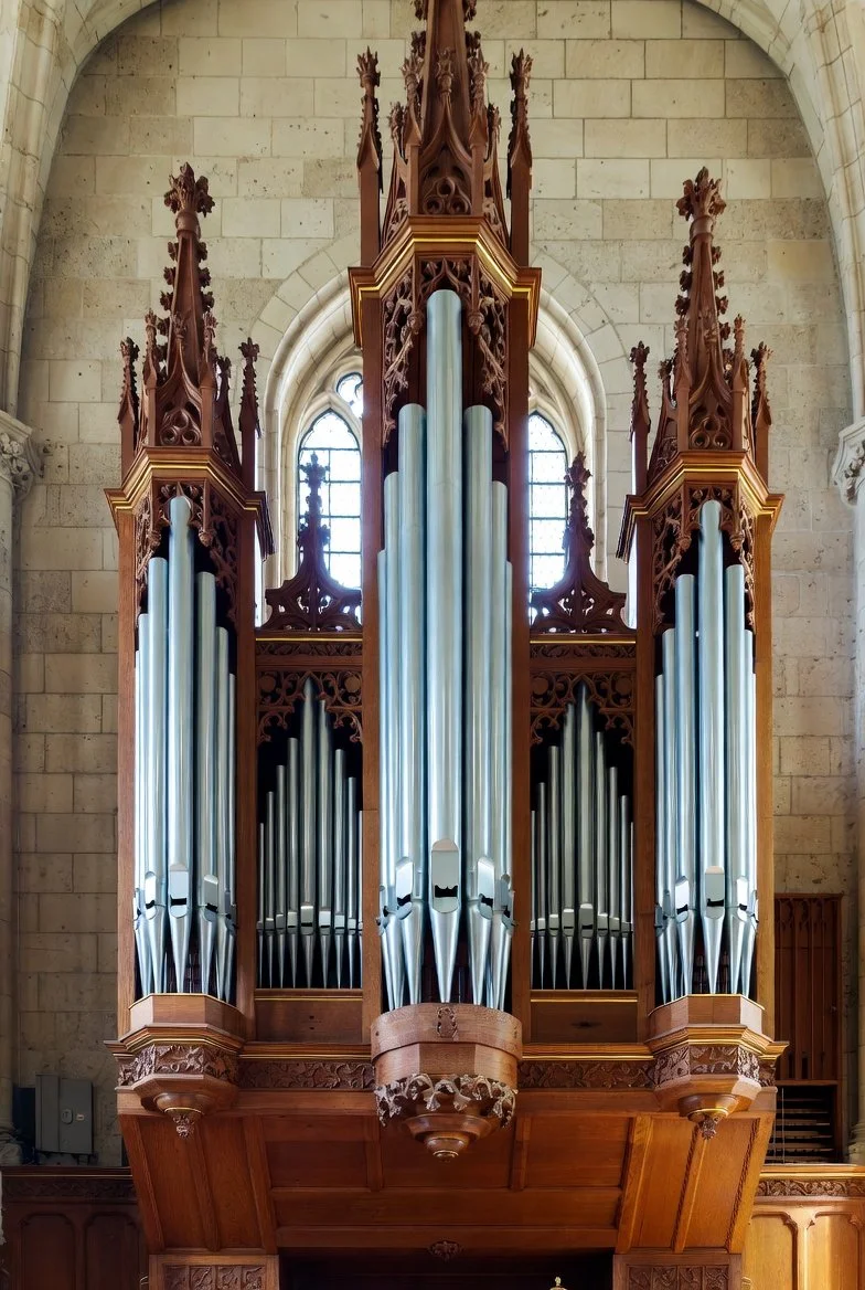 A large pipe organ with ornate wooden detailing inside a church with stone walls and arched windows.