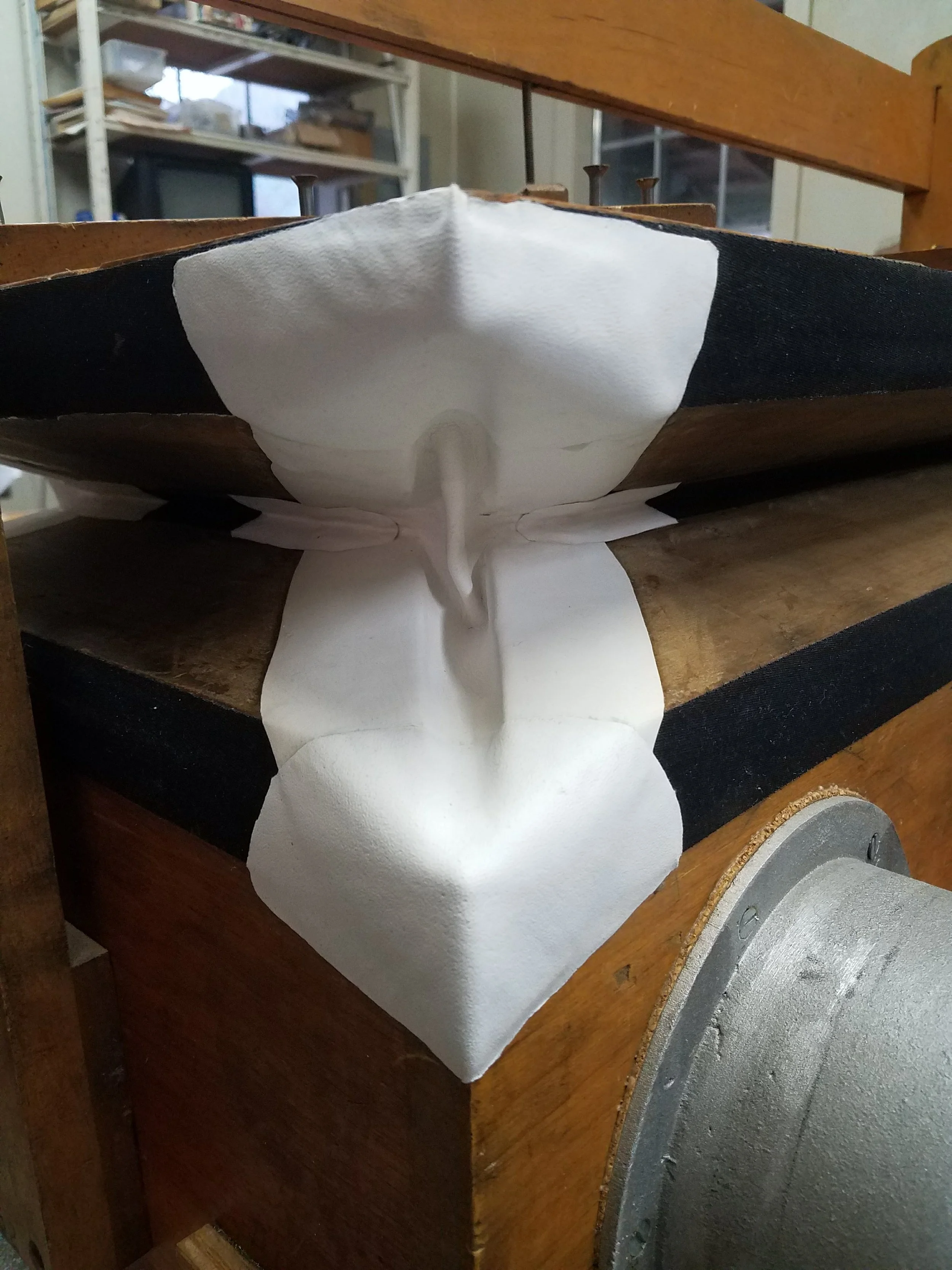 A white mask with black accents placed on a wooden surface, with part of a metal or plastic object visible, in a workshop or storage room. Shelves with boxes and equipment are in the background.