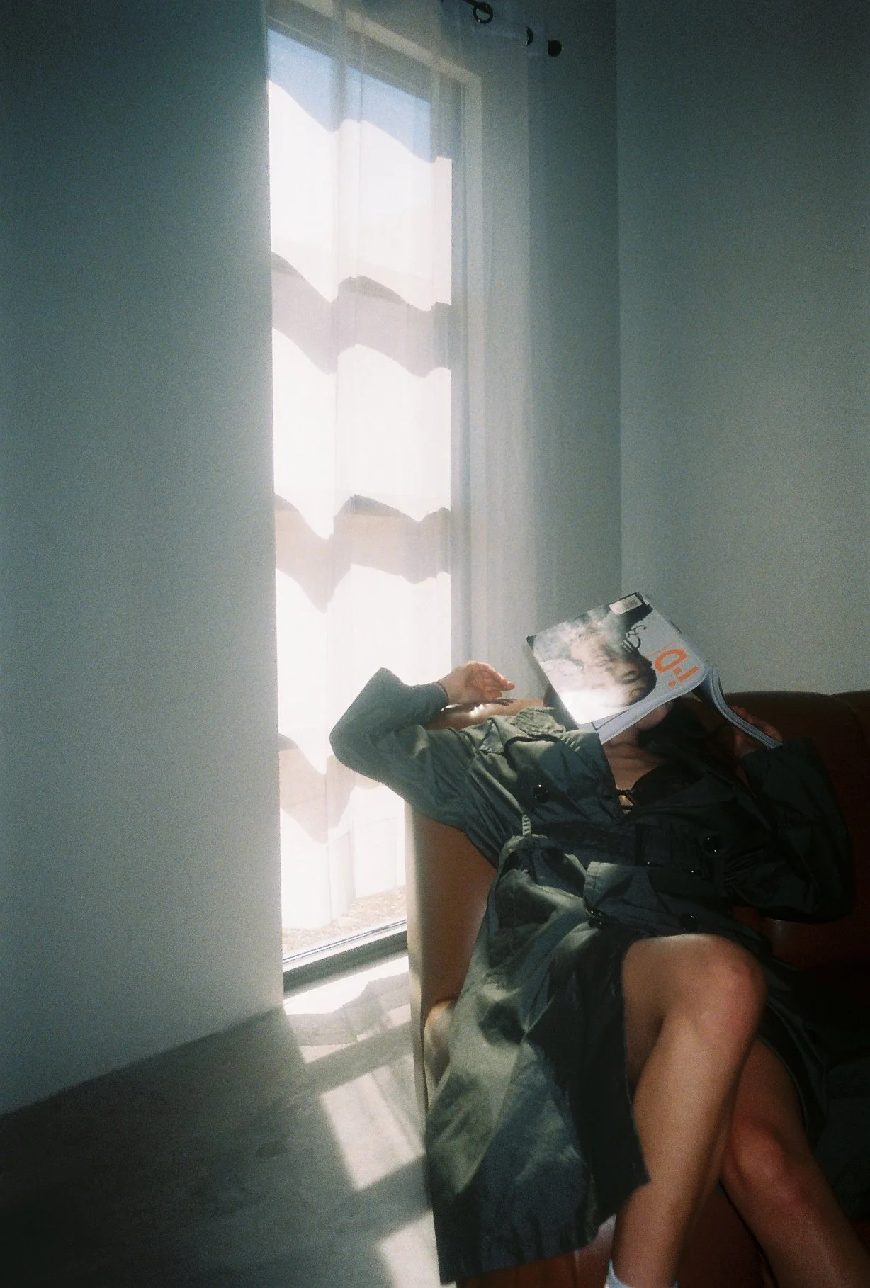 Person sitting on a sofa, reading a magazine, with sunlight streaming through a window behind them.