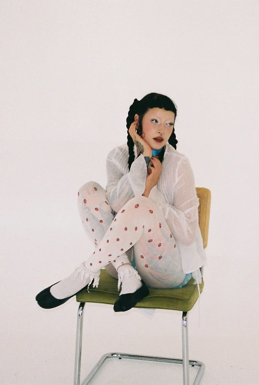 A young woman with black braided hair, white makeup, and colorful clothing, sitting on a green chair with a white background captured on 35mm film.