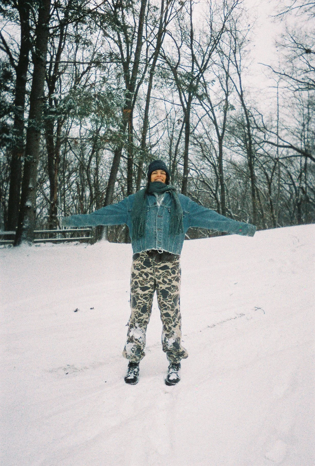 Person standing in snow with arms outstretched, wearing a denim jacket, camouflage pants, beanie, and scarf, in a winter forest setting.