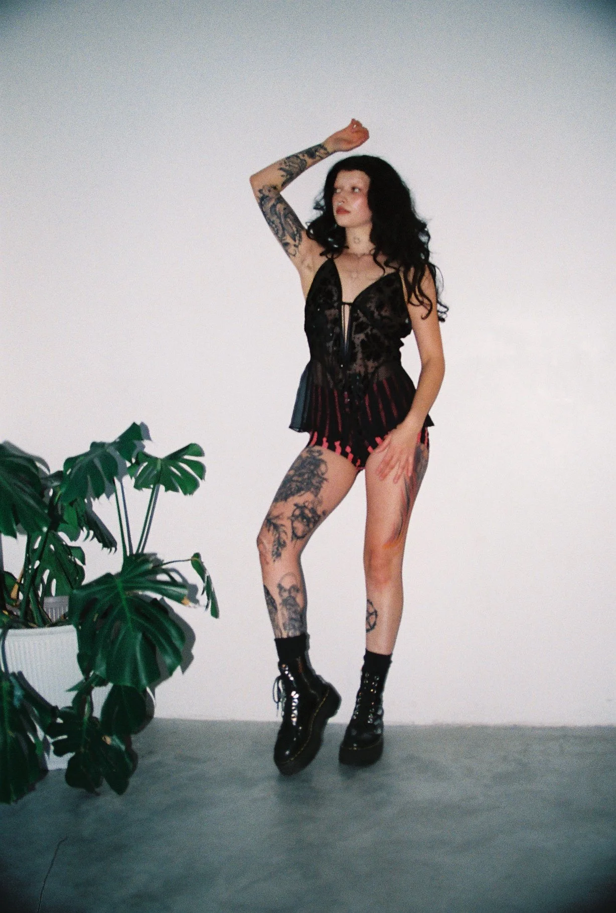 A woman with long black wavy hair and tattoos on her arms and legs, wearing a black lace corset top, red and black striped shorts, and black combat boots, standing against a plain white wall with a potted plant nearby.