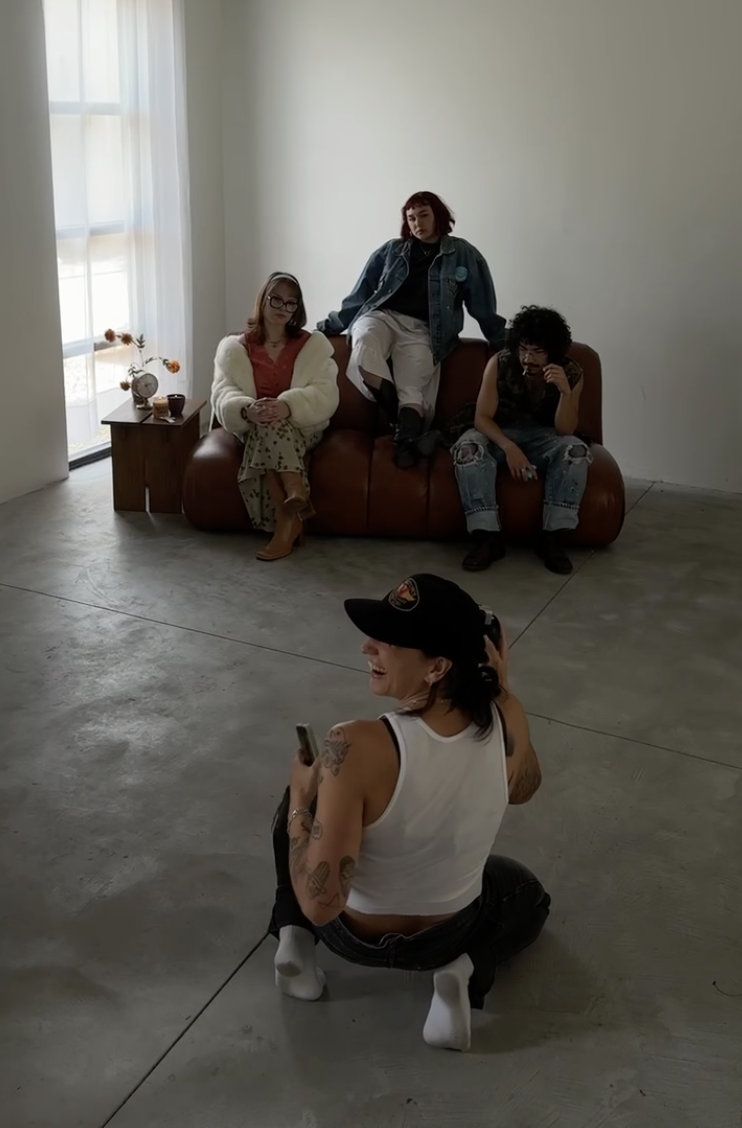 Four people in an art studio pose for photos. Three are sitting on a brown leather couch against a plain wall, and one is kneeling on the floor in front, holding a phone and smiling.