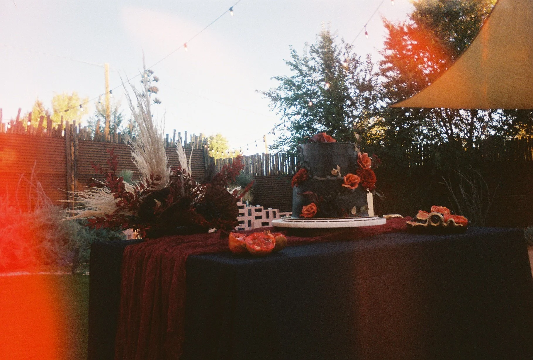 A decorated two-tiered black cake with orange and red flowers on top and around it, placed on a table with a dark tablecloth and burgundy runner, outdoors during daylight with trees and a wooden fence in the background.
