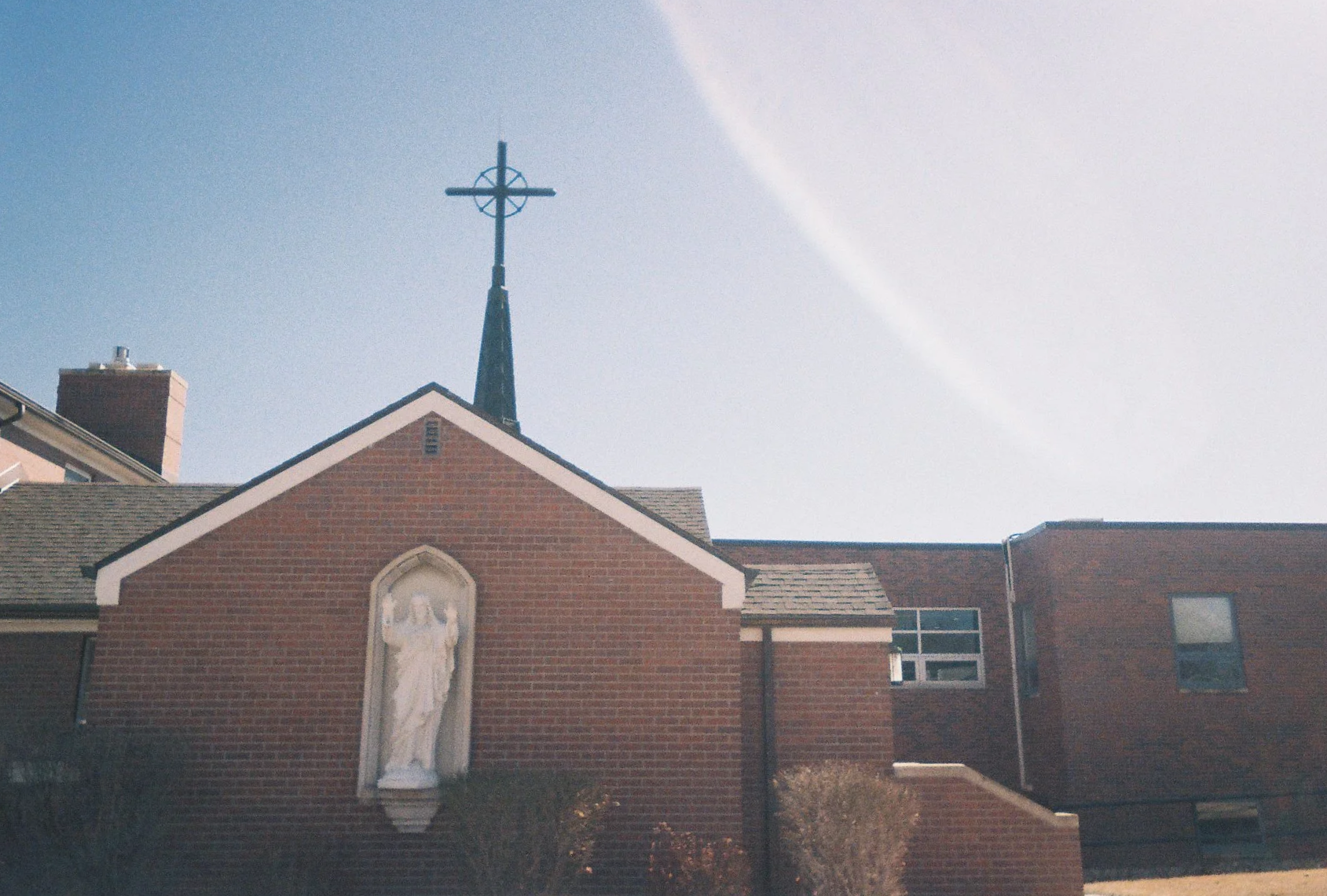 A brick church with a steeple topped by a cross, and a stone religious statue near the front.