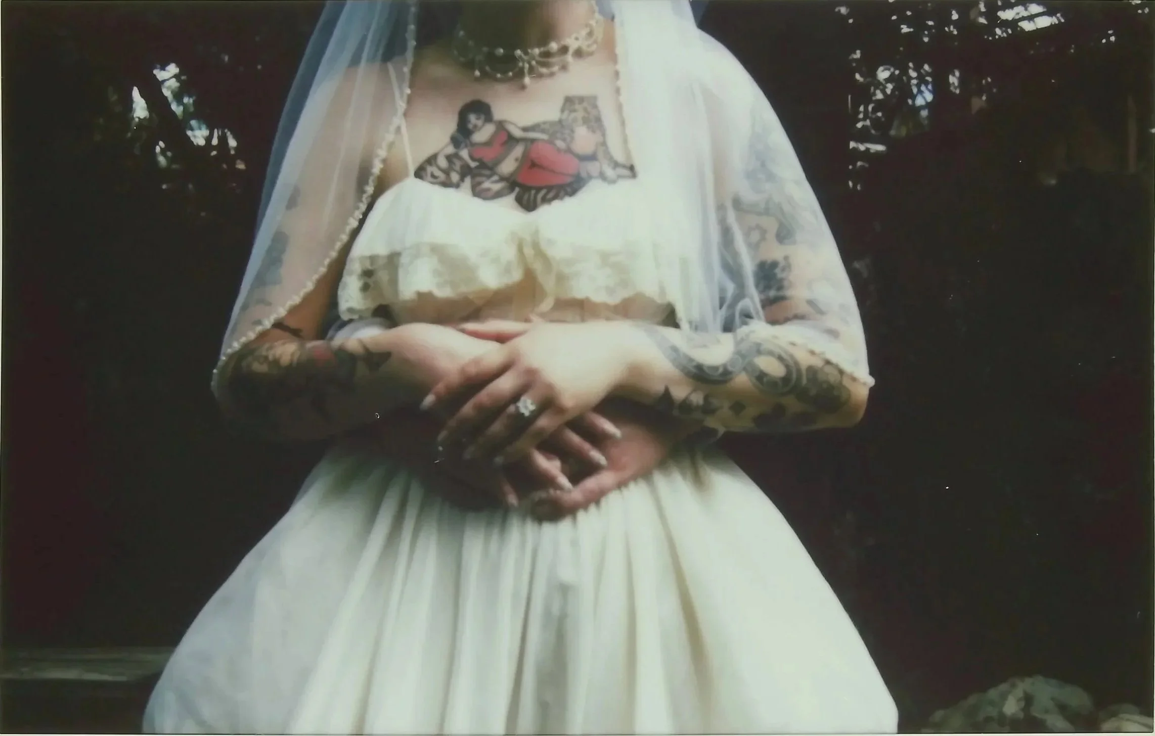A bride wearing a white wedding dress with lace sleeves and intricate patterns. She has tattoos on her arms and a tattoo of a woman in a red dress on her chest. The bride is holding her hands together in front of her. She is wearing a veil and a pearl necklace. The background is dark with trees visible.