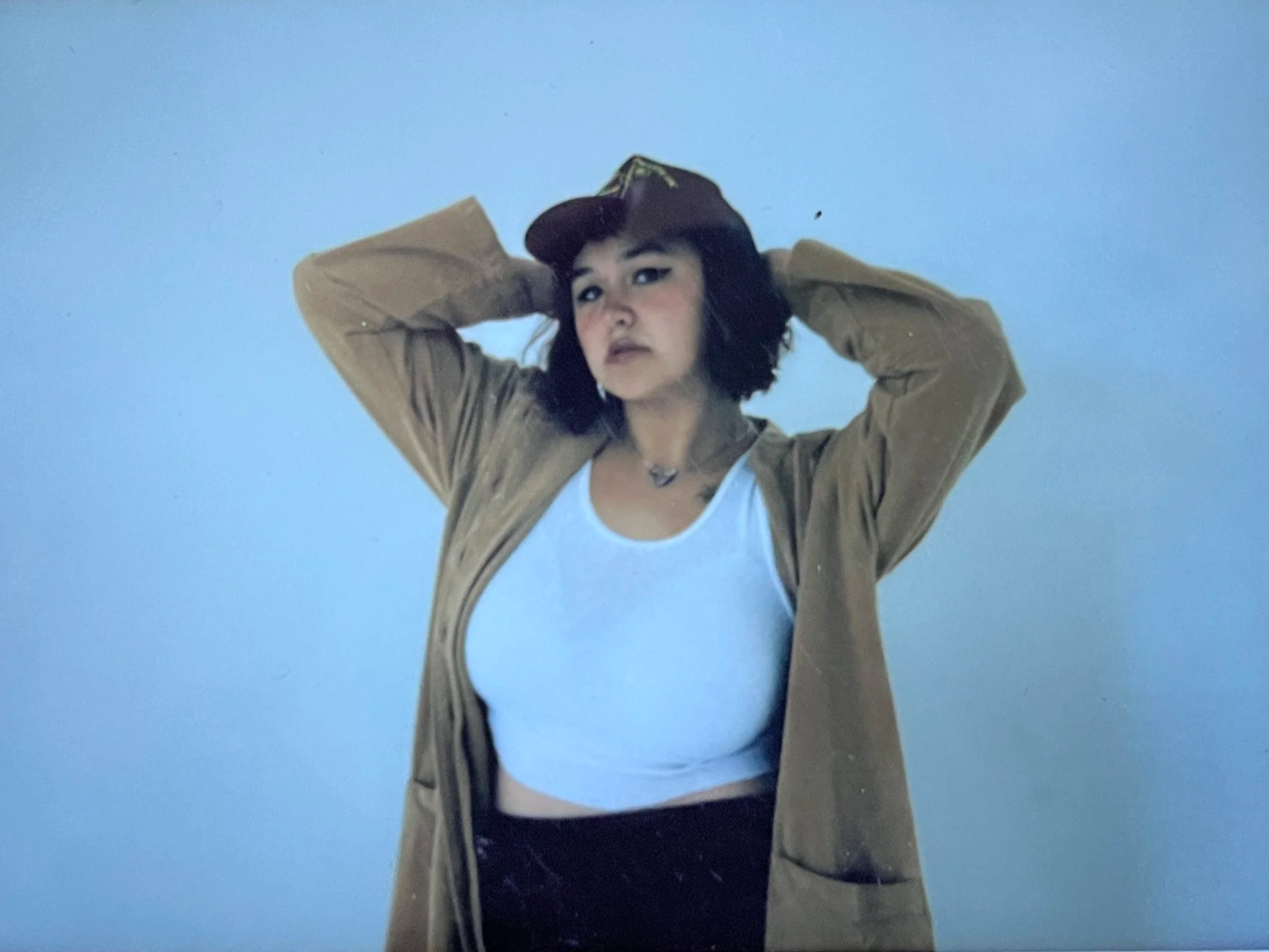 A woman with short dark hair wearing a beige jacket, white crop top, and a cap, standing against a plain light blue background, looking slightly to the side, with her hands behind her head.
