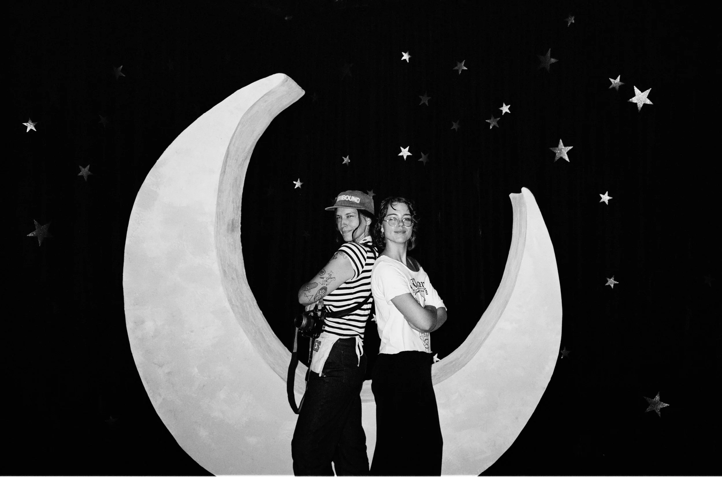 Two women standing in front of a moon-shaped cutout and a starry background, posing for a photo. One woman has glasses and curly hair, while the other is wearing a cap, striped shirt, and tattoos.