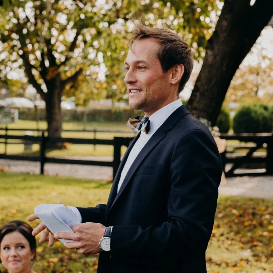 A man in a suit and bow tie holding papers, smiling outside with a woman nearby, trees, and a wooden fence in the background.

Mann im Anzug bei einer Traurede