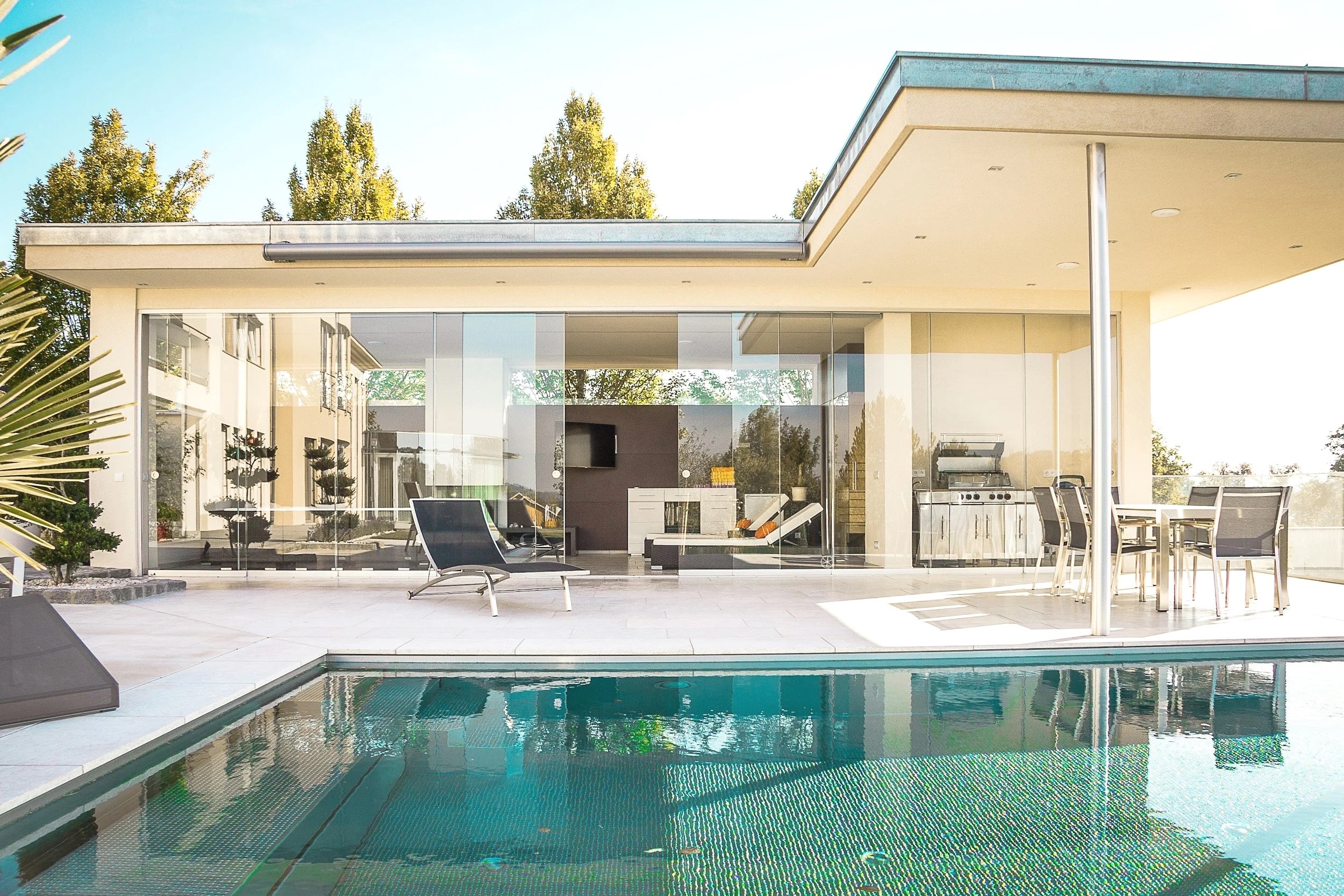 Modern house with a glass backyard patio, outdoor furniture, a swimming pool, and tall trees in the background.