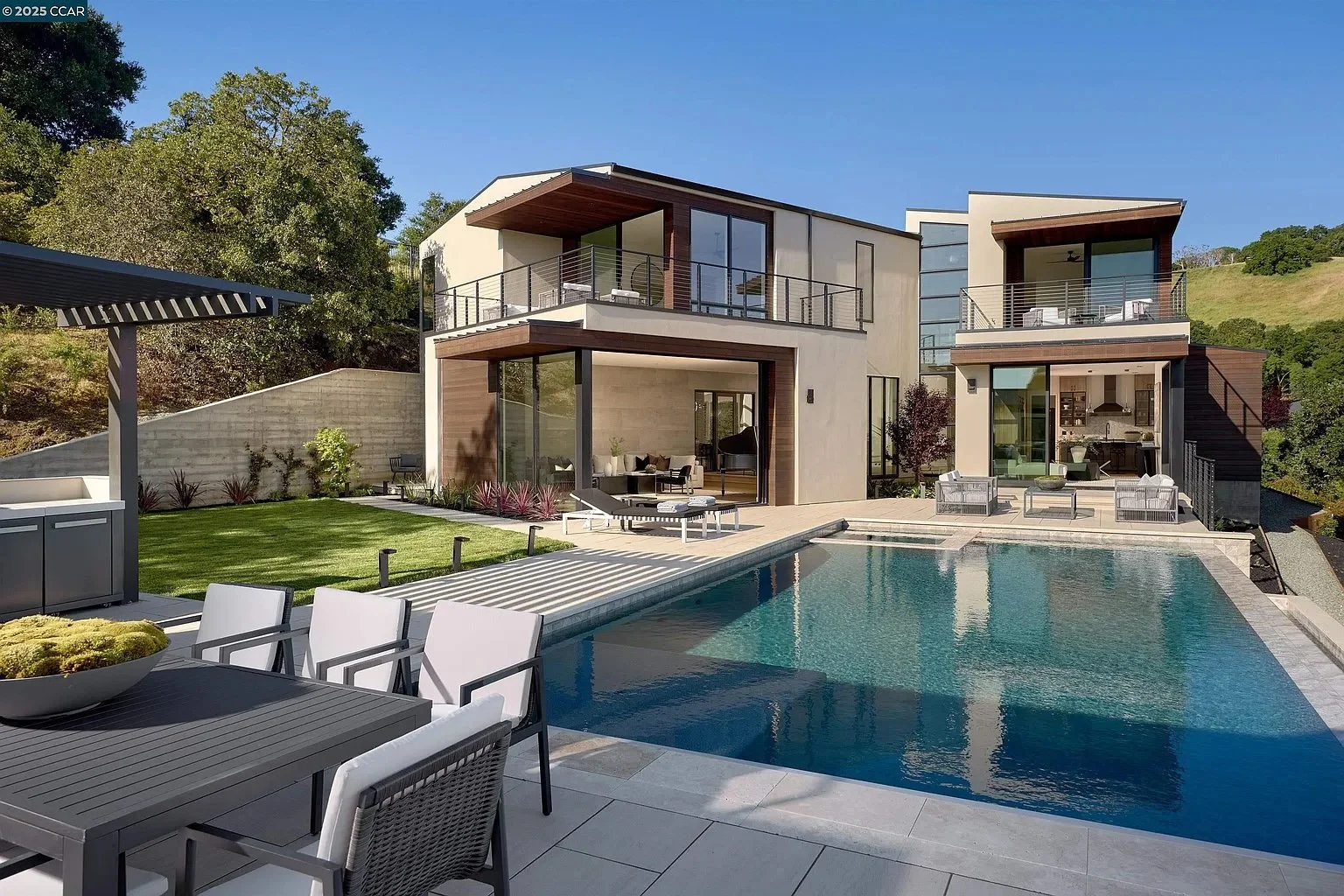 Modern multi-level house with large windows and balconies, backyard with swimming pool, outdoor seating, and well-maintained lawn, surrounded by trees and hillside.