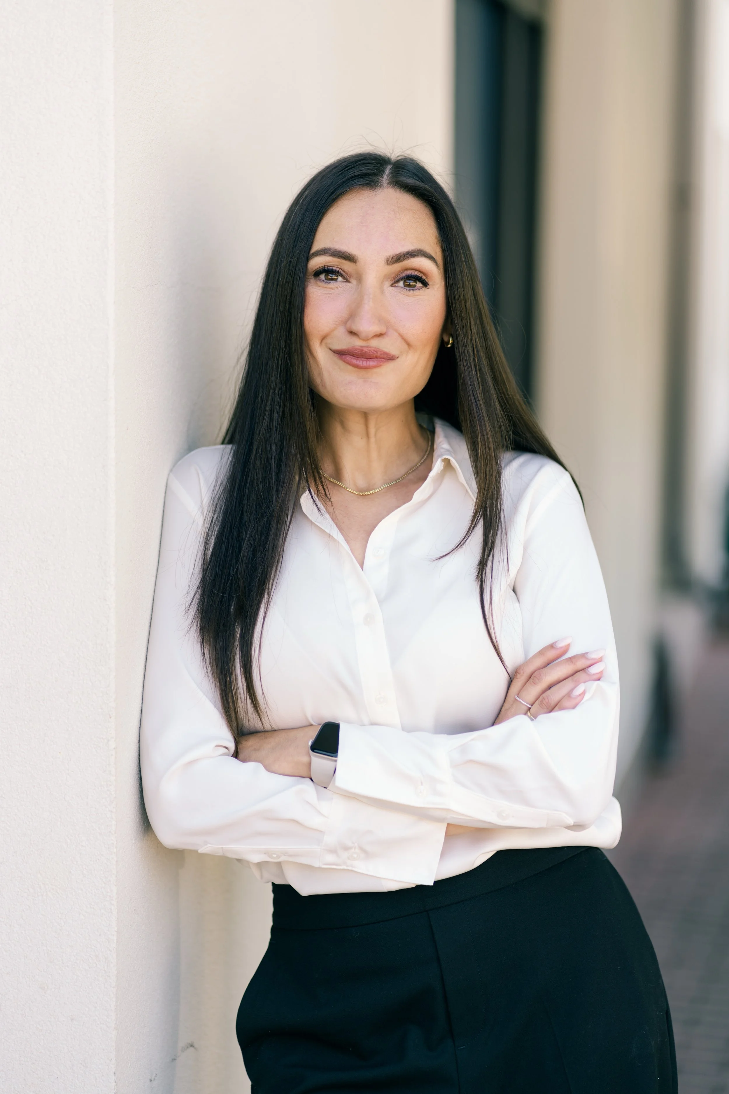 A woman with long dark hair, wearing a white blouse, black skirt, and a smartwatch, standing with arms crossed against a beige wall, smiling at the camera.