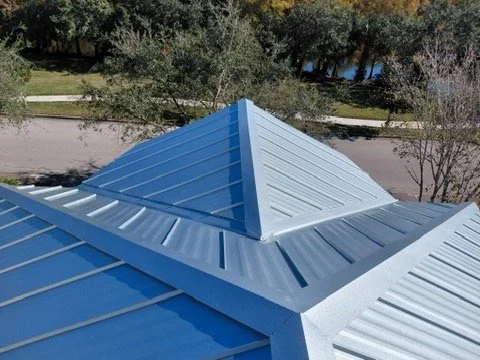 Close-up of a metal roof with a prominent flashing peak, showing blue, white, and gray panels, with trees in the background.
