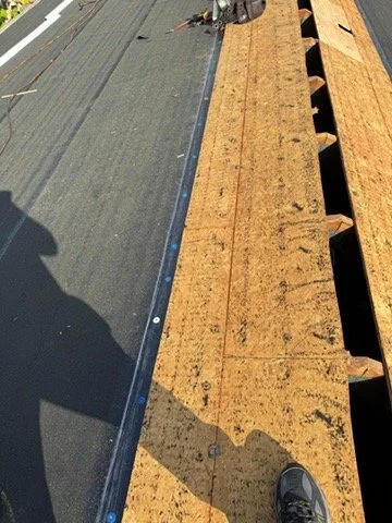 Part of a roof under construction showing wooden framing, a black protective layer, and part of a person's shadow and shoe.