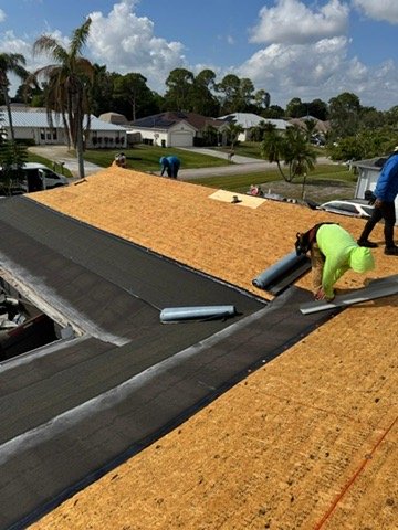 Workers installing new roofing on a house, with some sheets of roofing material already in place, in a neighborhood with trees and houses in the background.