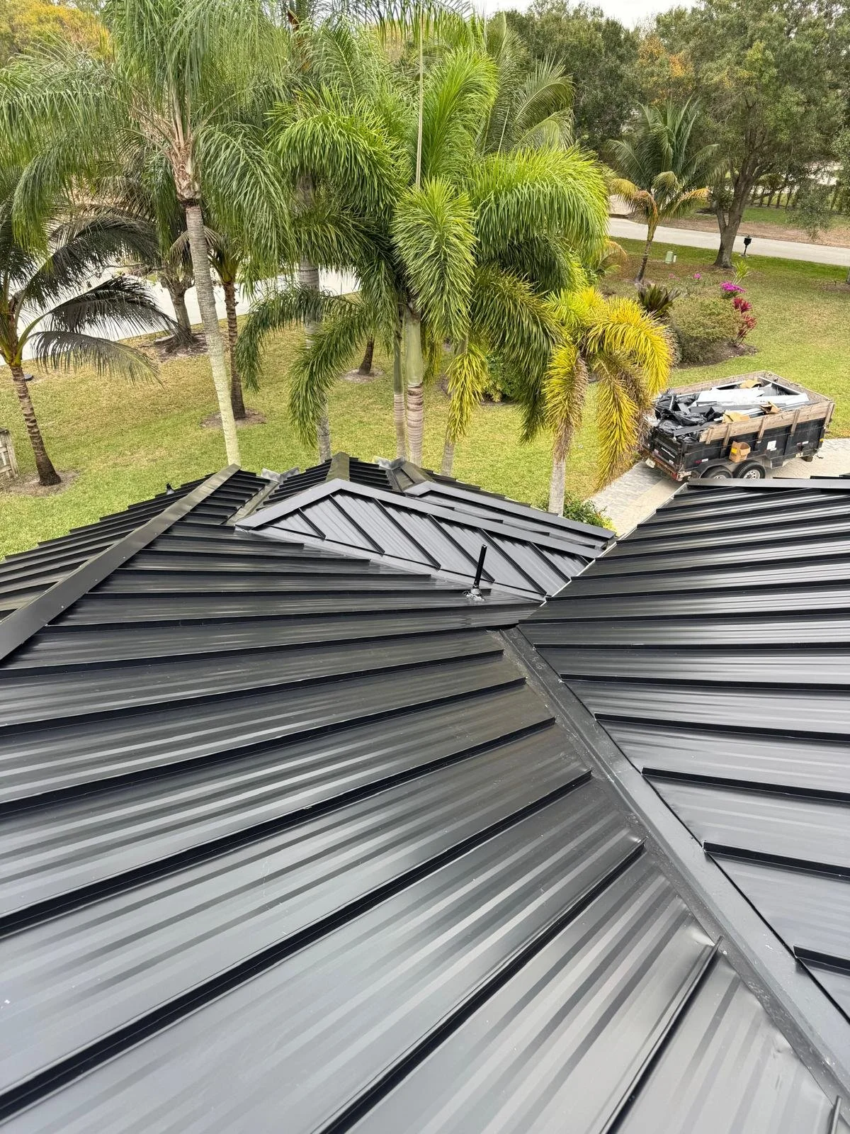 View of a metal roof with multiple pitches, surrounded by palm trees and greenery, with a black utility trailer parked on a driveway in the background.