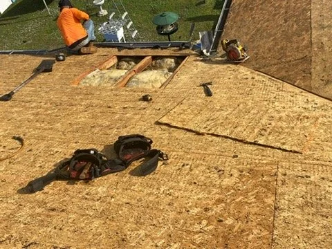 Construction worker installing roof sheathing on a building, with roofing tools and equipment nearby.
