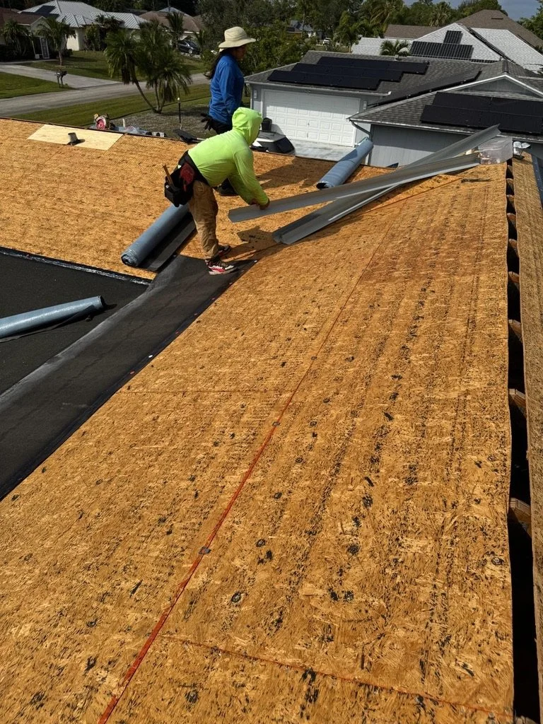 Two construction workers installing roofing material on a house, with solar panels installed on neighboring rooftops in a suburban neighborhood.