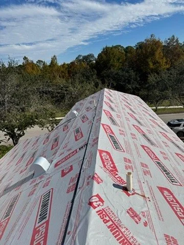 The roof of a building under construction covered with weather barrier sheathing, with a downspout and small vent pipe visible.
