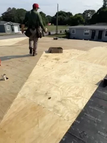 A construction worker walking on a flat roof with plywood sheets, holding tools, with a small building and trees in the background.