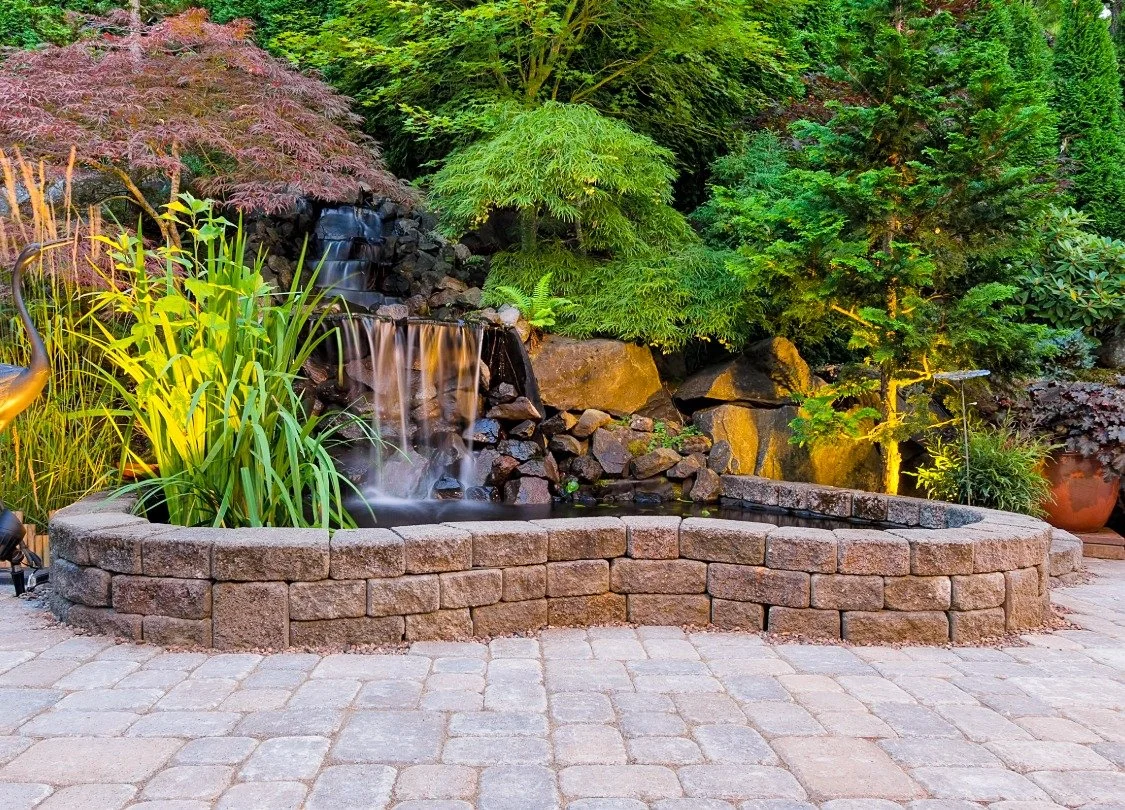 Cascading natural stone water feature design with custom retaining wall, pavers surrounded by plants and trees in Bend