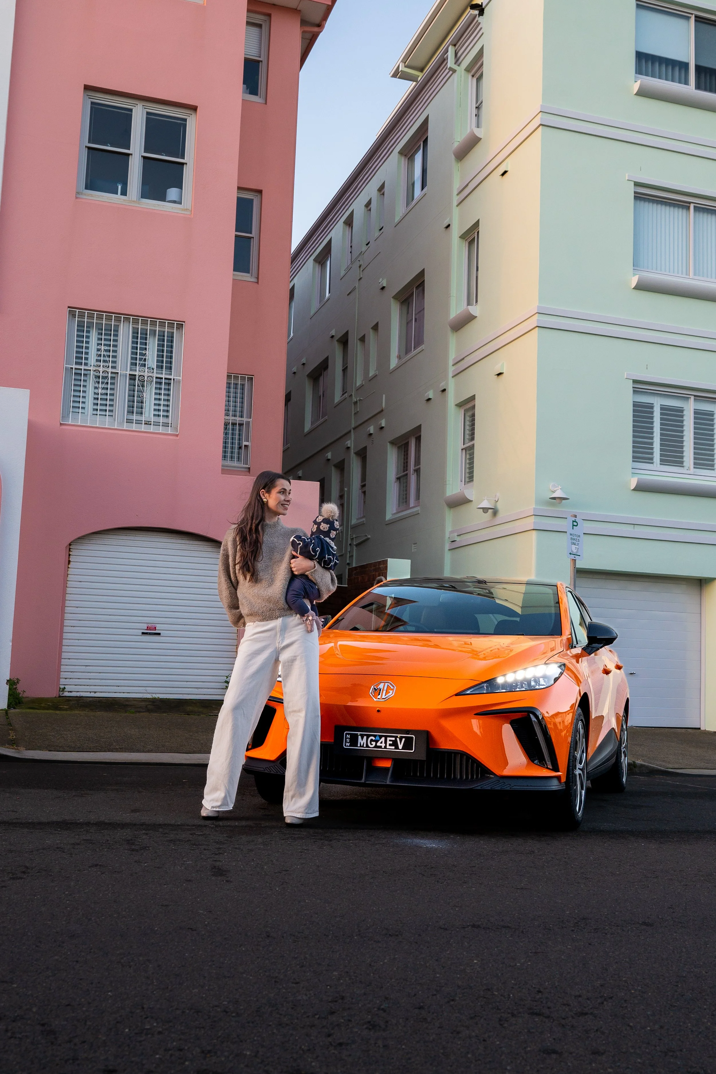 A woman holding a child standing next to an orange MG electric car parked on a city street with pastel-colored apartment buildings in the background.
