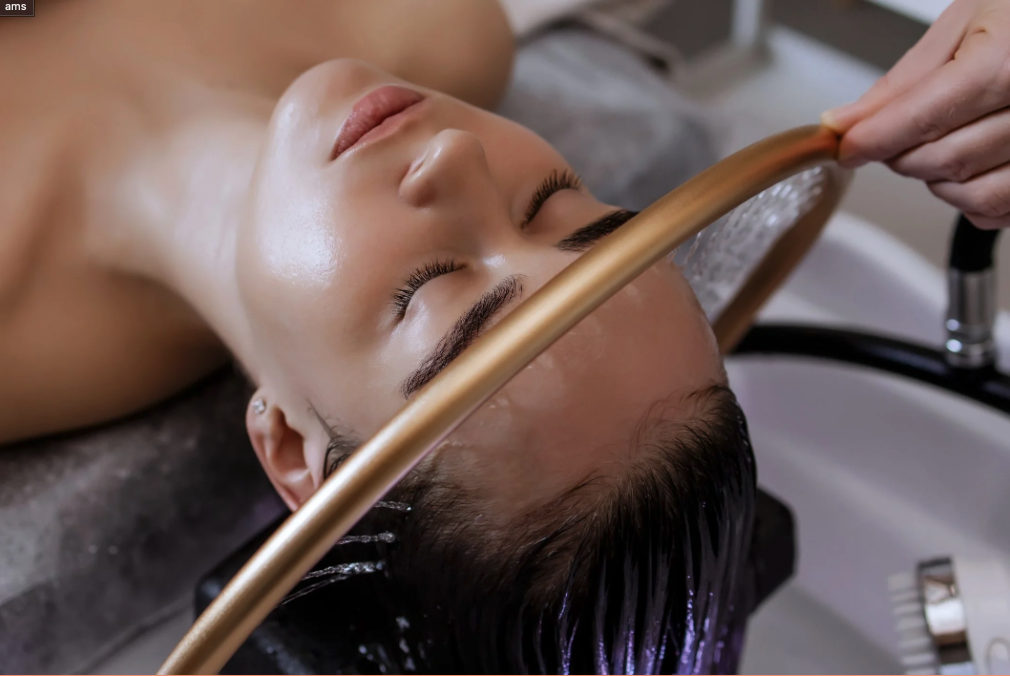 A woman receives a haircut treatment or scalp cleaning at a salon, lying back with eyes closed under a hair washing station. Waterfall Faucet used for A.M. Head Spa Treatments