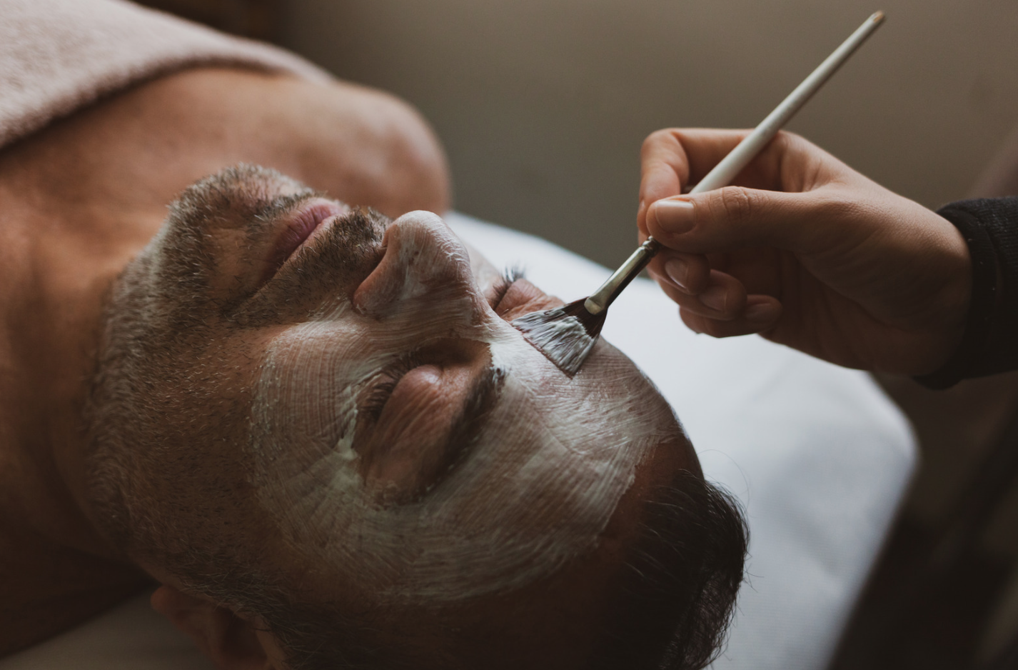 A man lying down with a facial mask being applied by a person using a brush.