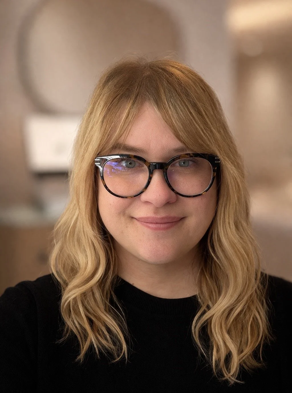 A woman with wavy blonde hair and glasses smiling indoors.