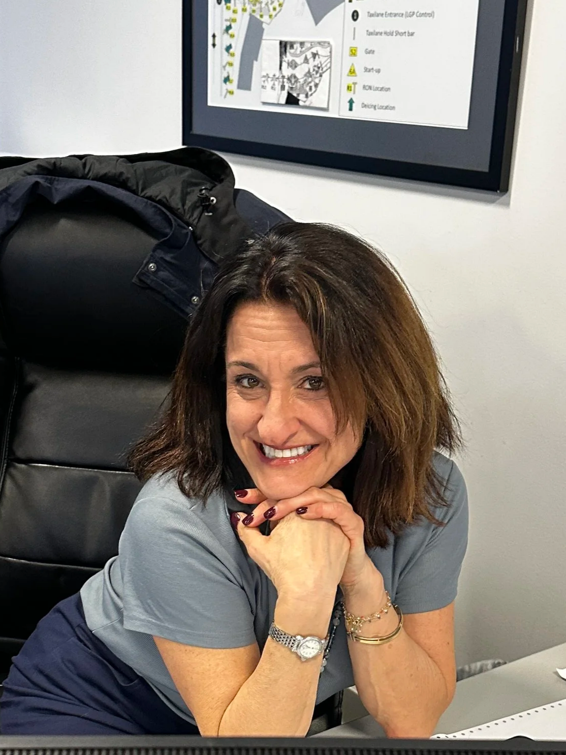 Headshot of a woman with medium-length brown hair, wearing a black blazer and a white top, smiling at the camera against a grey background.