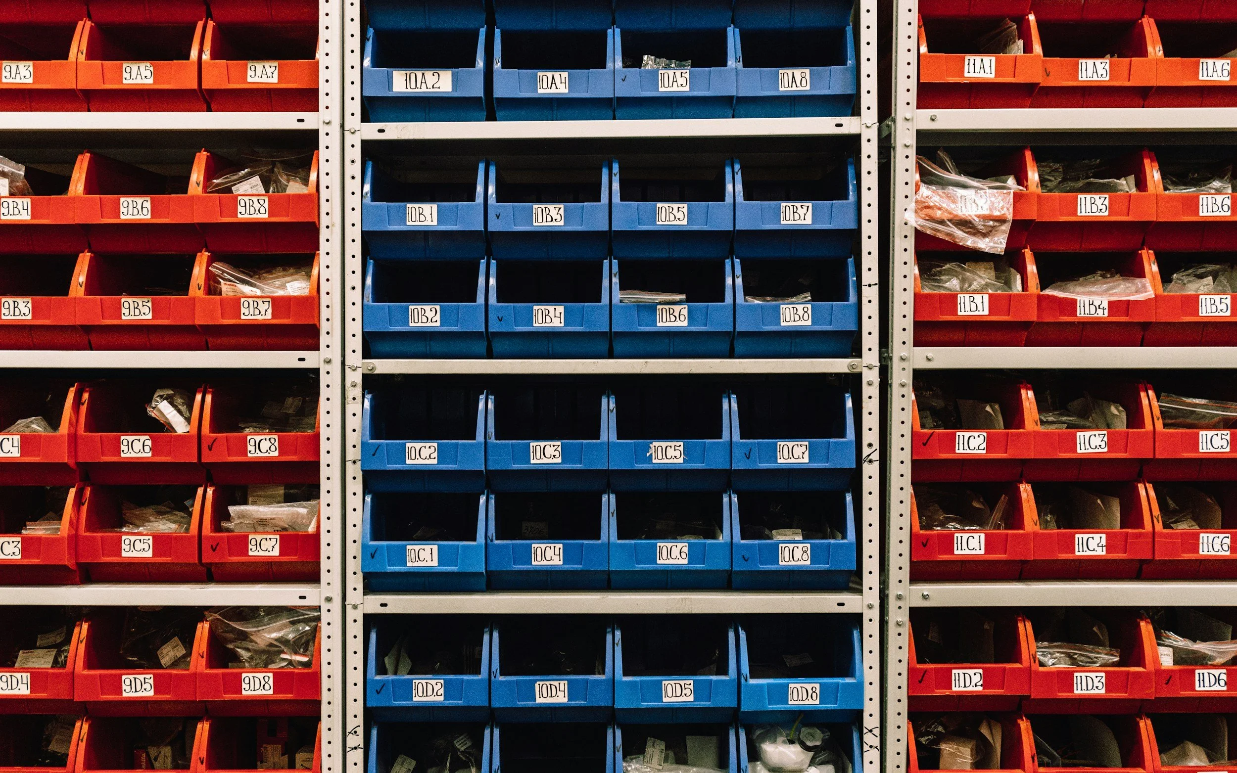 A large storage rack with multiple orange and blue plastic bins, each labeled with alphanumeric tags, filled with various small hardware items in a warehouse or workshop.