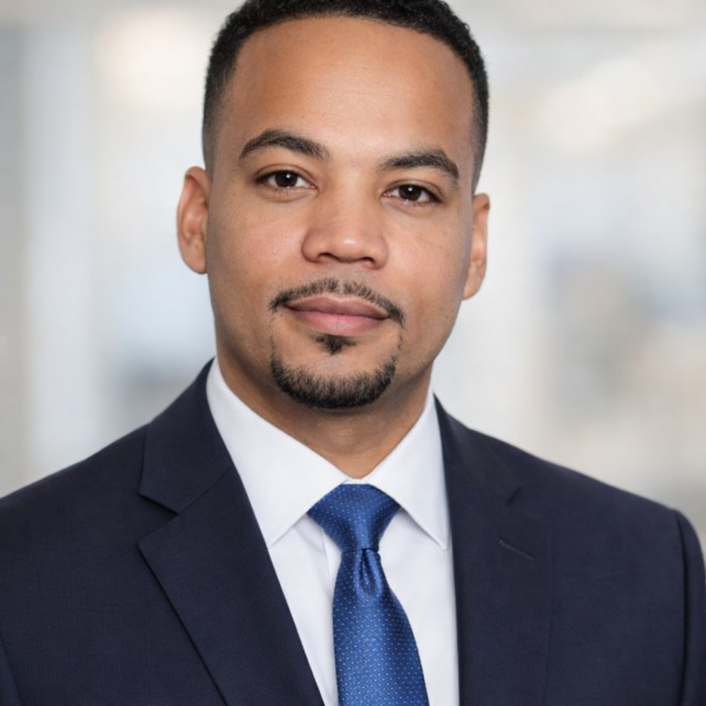 Professional headshot of a man in a dark suit, white shirt, and blue tie, with short black hair and a goatee, in an office setting.