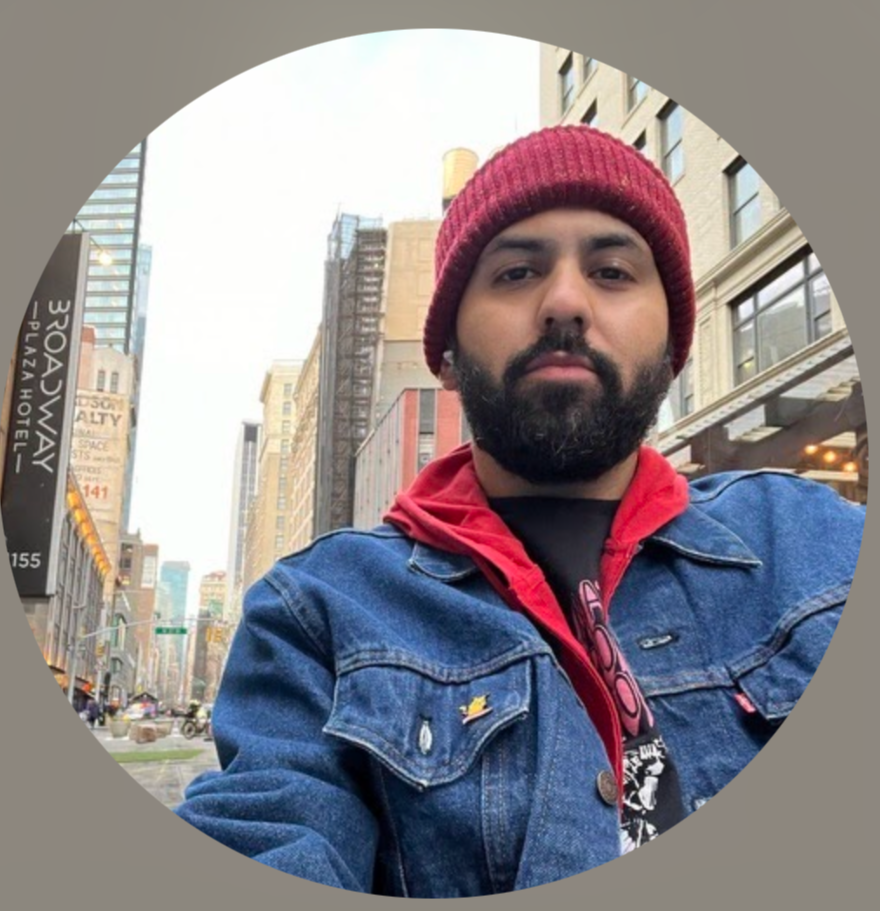 Headshot of a smiling African American man in a light gray suit, light blue shirt, and dark blue tie against a dark background.