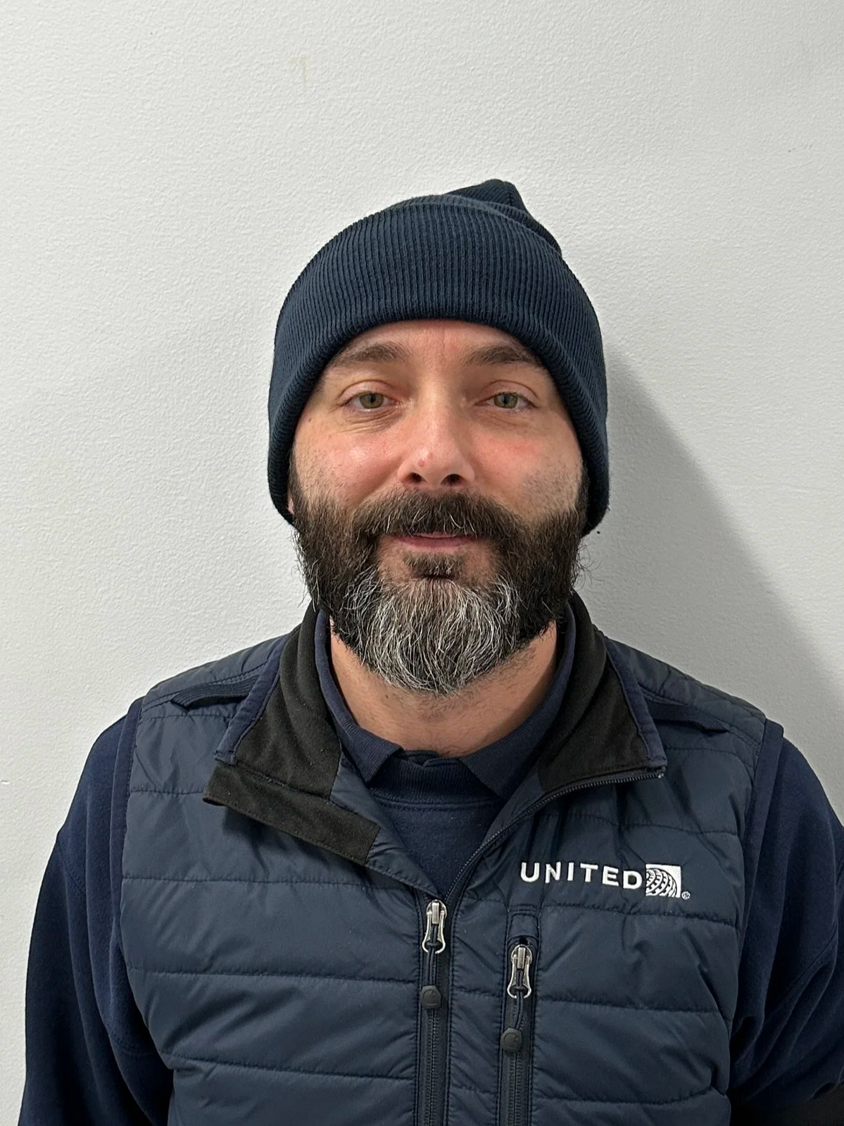 Headshot of a man with a bald head, beard, and mustache wearing a dark suit, white shirt, and dark tie against a plain background.