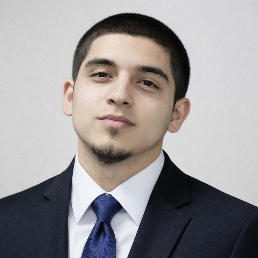 A young man in a business suit with a white shirt and blue tie, posing against a plain light gray background.