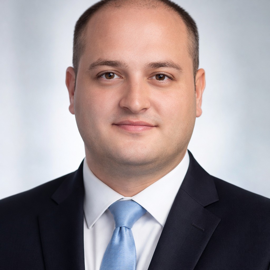 Headshot of a man in a suit with a light blue tie, smiling slightly, with a neutral background.