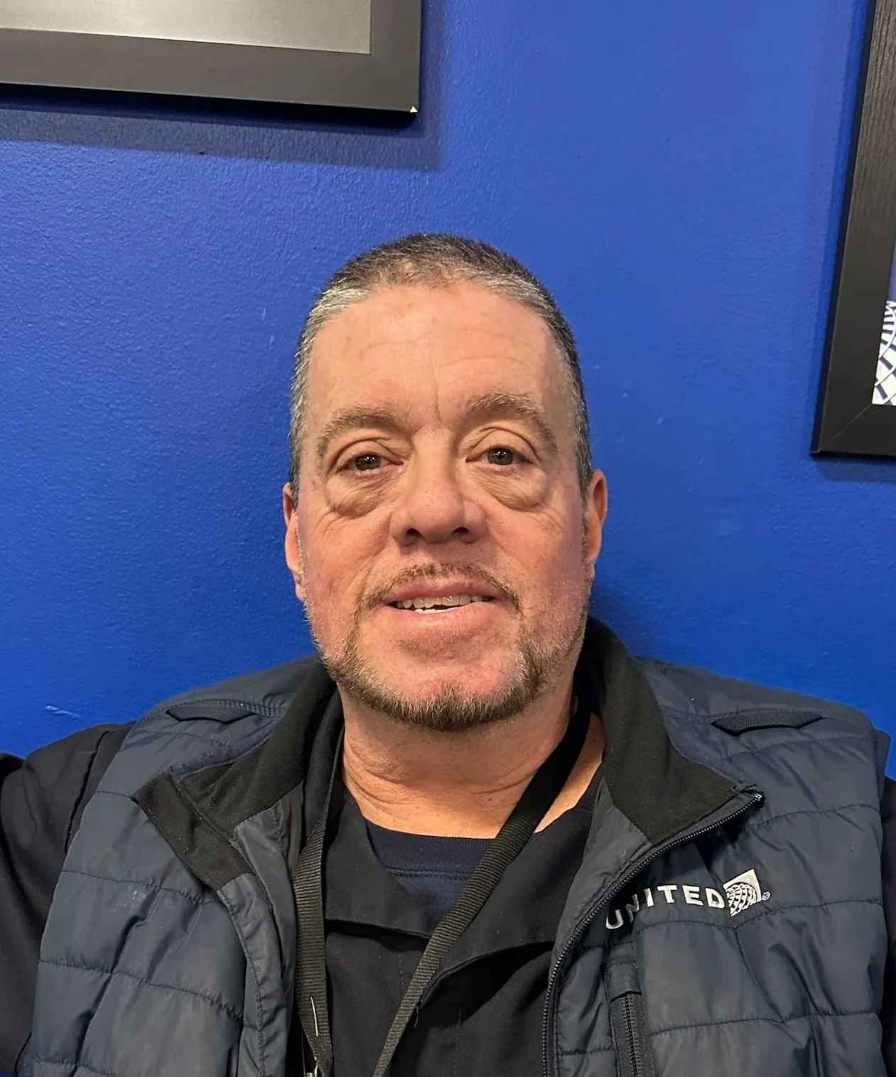 A man with short hair, facial hair, wearing a black jacket and a lanyard, seated against a blue wall.