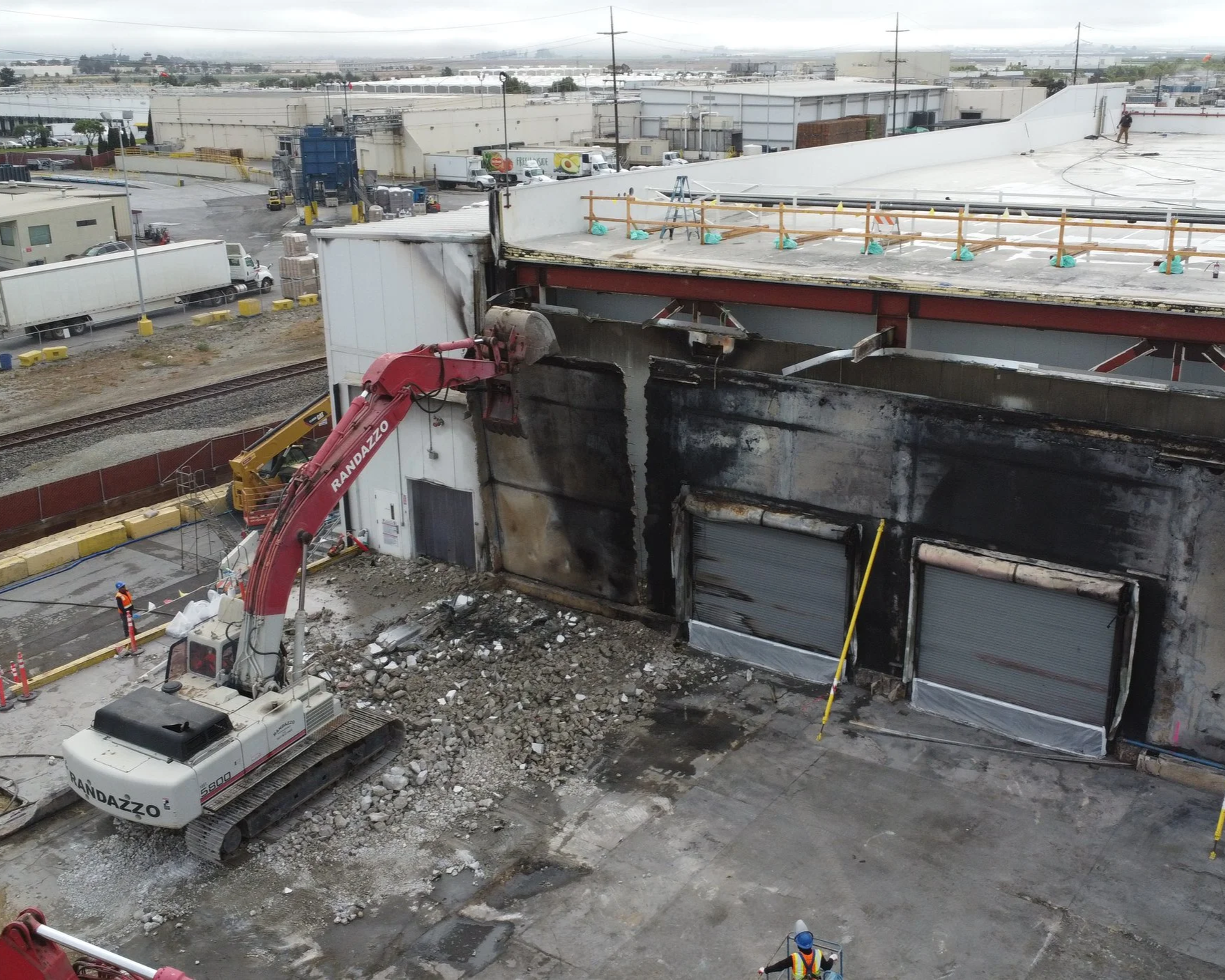 Construction workers and an excavator demolishing or working on the concrete structure of a large industrial building or warehouse with burned and damaged exterior walls.