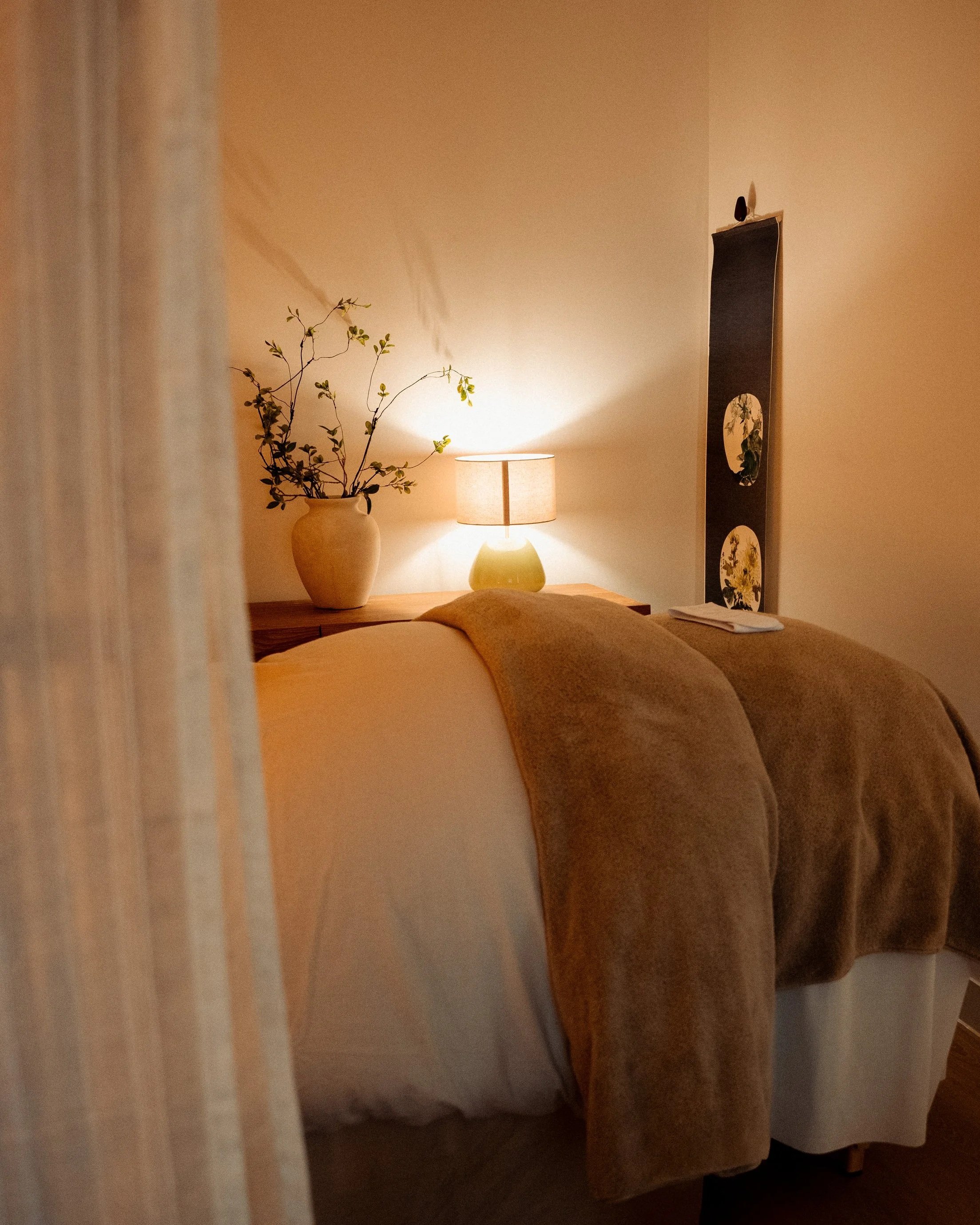A cozy bedroom with a bed covered in brown blankets, a bedside table with a lamp, a decorative vase with branches, and framed artwork on the wall.