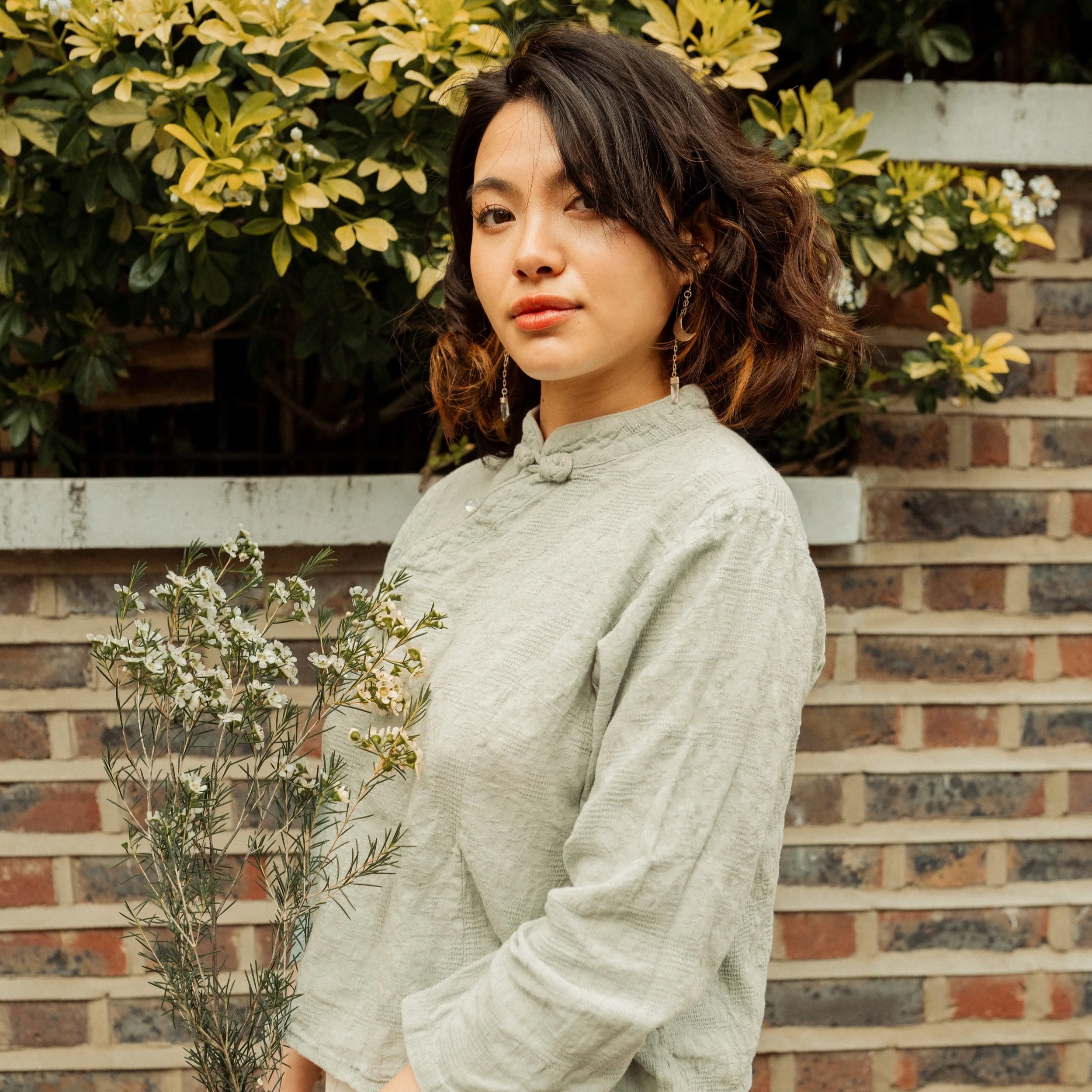 A woman with shoulder-length wavy hair holding a small bouquet of white flowers, standing outdoors in front of a brick wall and green bushes.