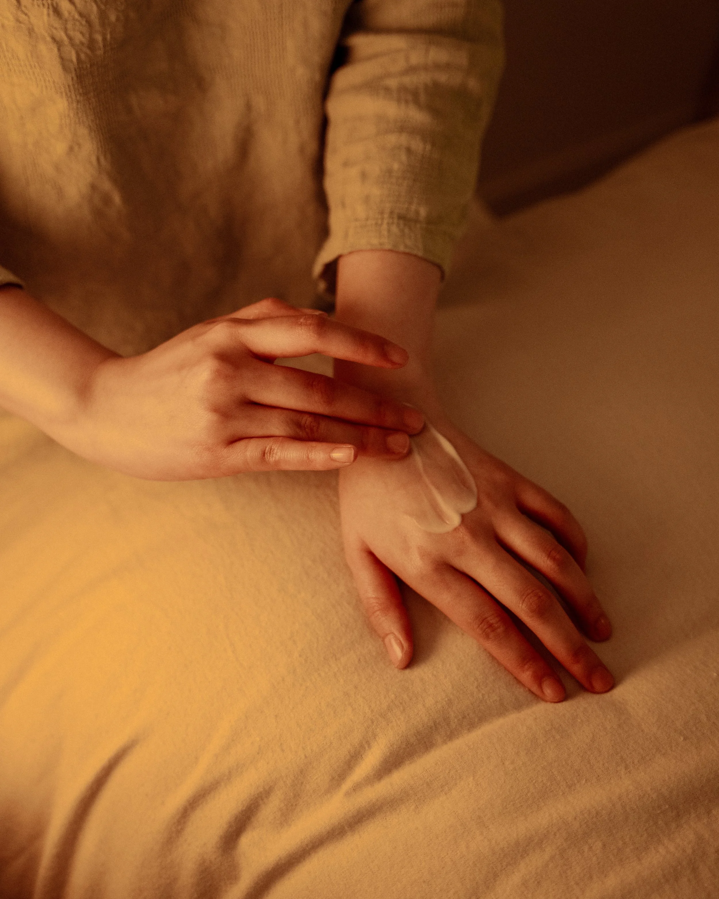 Close-up of hands with one hand gently resting on top of the other on a soft, beige surface, with warm lighting.