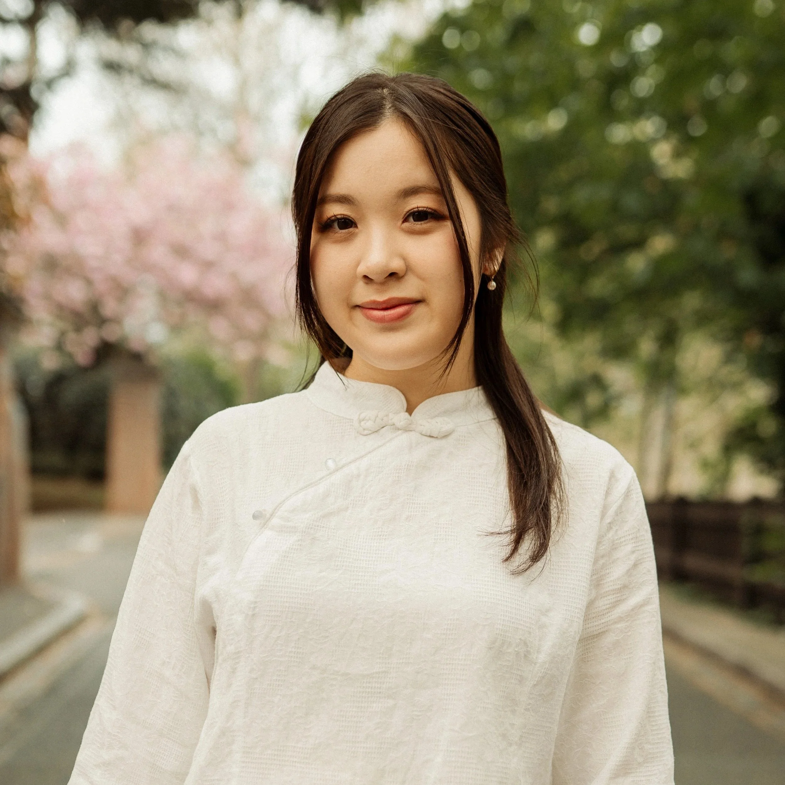 A young woman standing outdoors with trees and pink blossoms in the background, wearing a white traditional Chinese-style top.