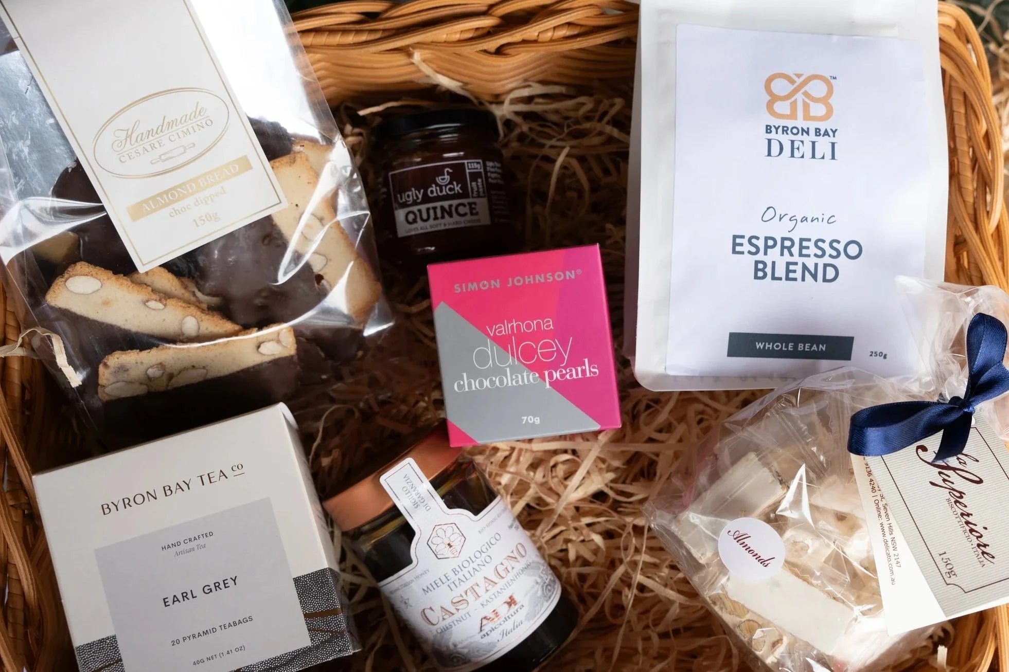 A gift basket containing almond biscotti, a jar of ugly duck quince jam, a box of Valrhona Dulcey chocolate pearls, a bag of Byron Bay Organic Espresso Blend coffee, Earl Grey tea, a bottle of Castagno organic maple syrup, and almond-flavored macaron