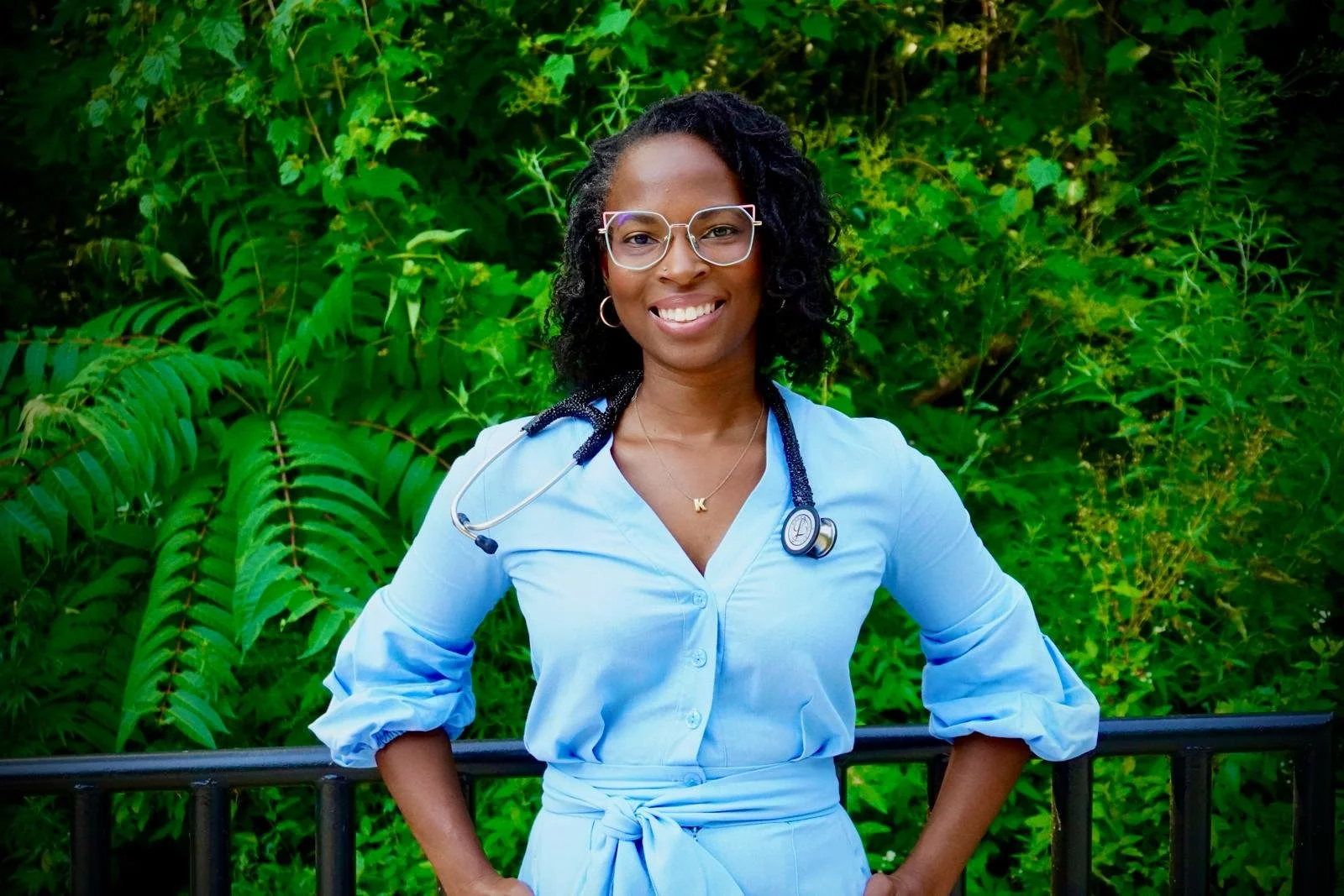 A smiling African American female healthcare professional standing outdoors in front of green foliage, wearing medical scrubs, glasses, and a stethoscope around her neck.