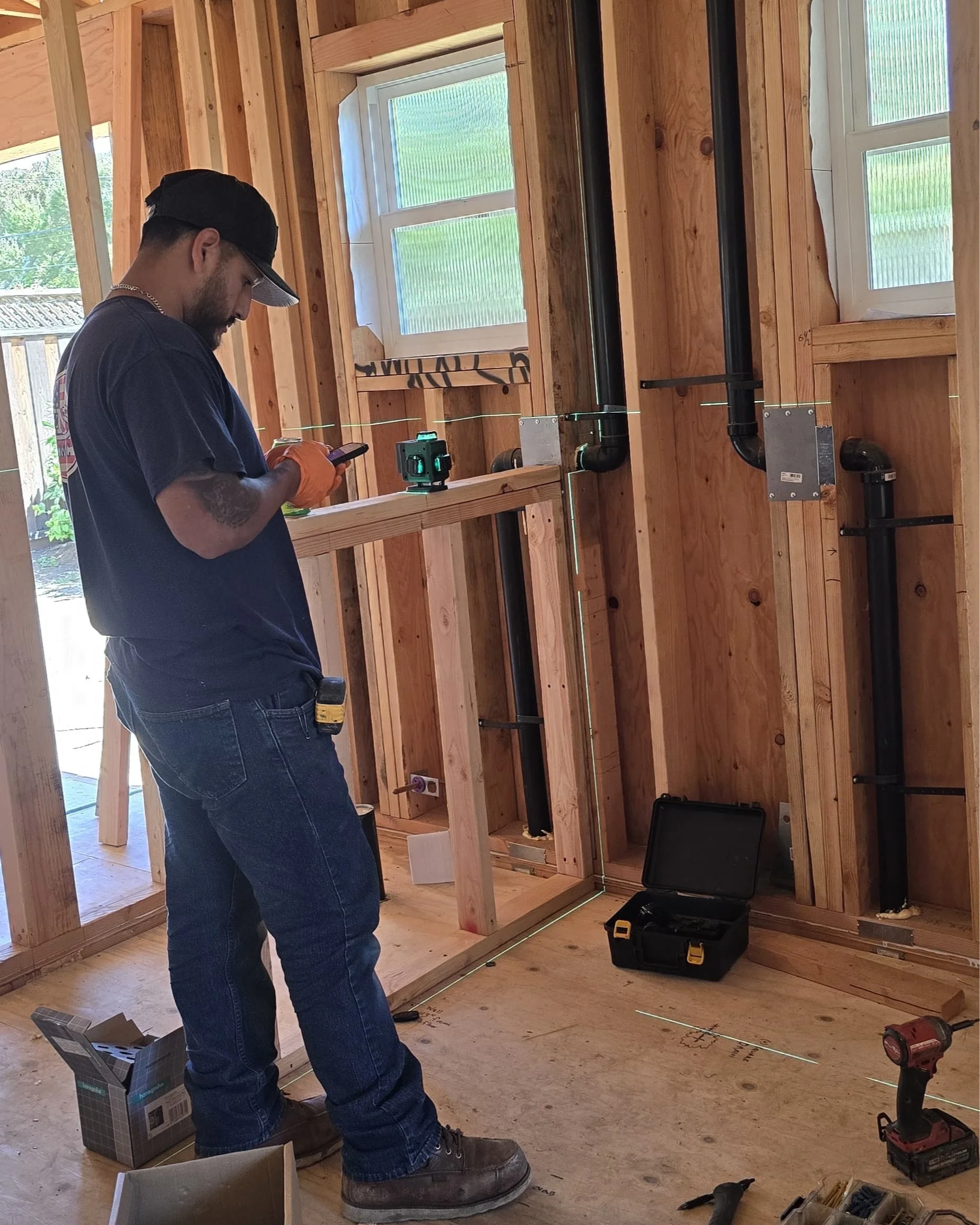 A man working on a construction site, standing inside a wooden framed room, using a measuring tool, with electrical wiring and piping installed on the walls, and construction tools and materials around.